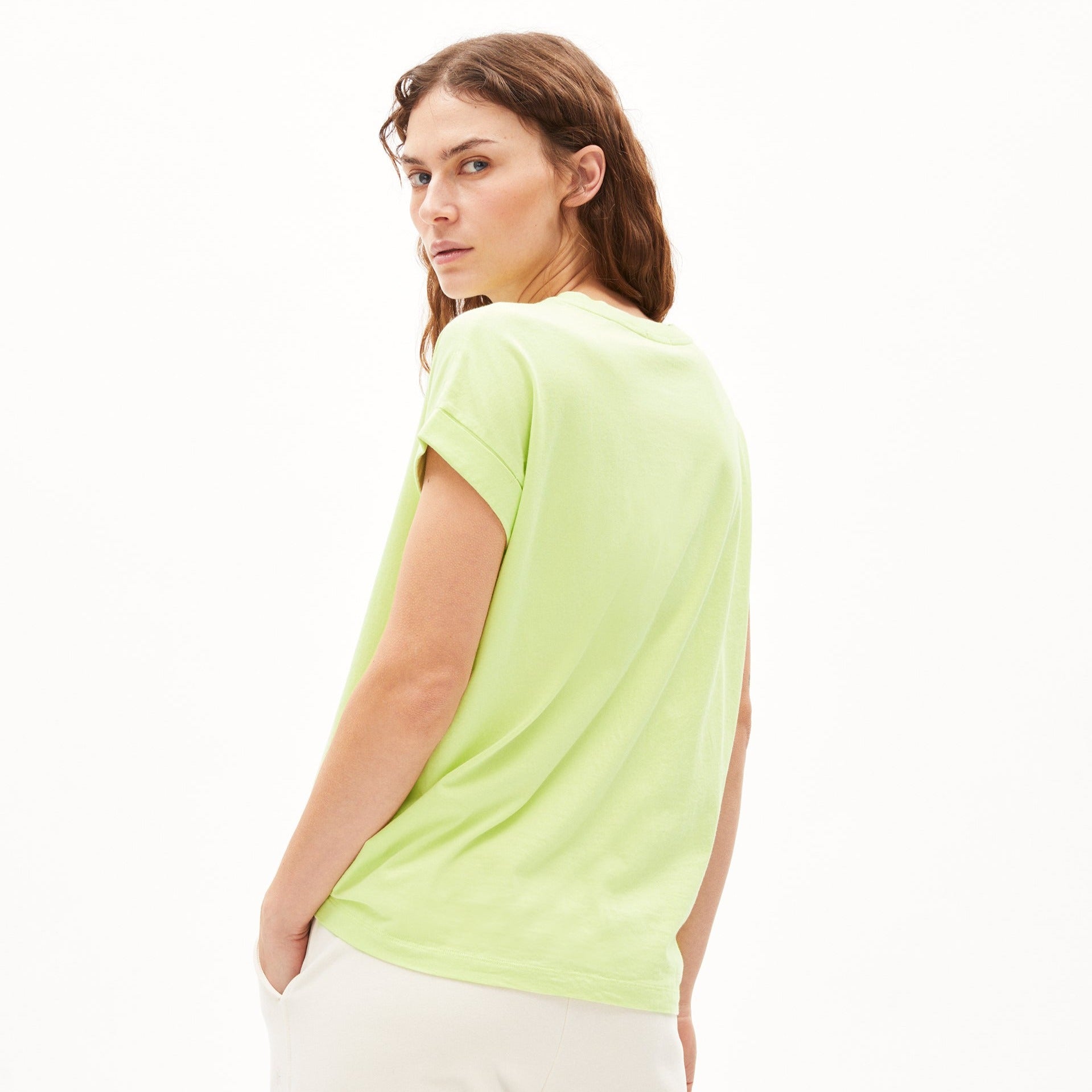 Person mit langen Haaren trägt ein hellgrünes T-Shirt und steht seitlich, während sie über die Schulter schaut.