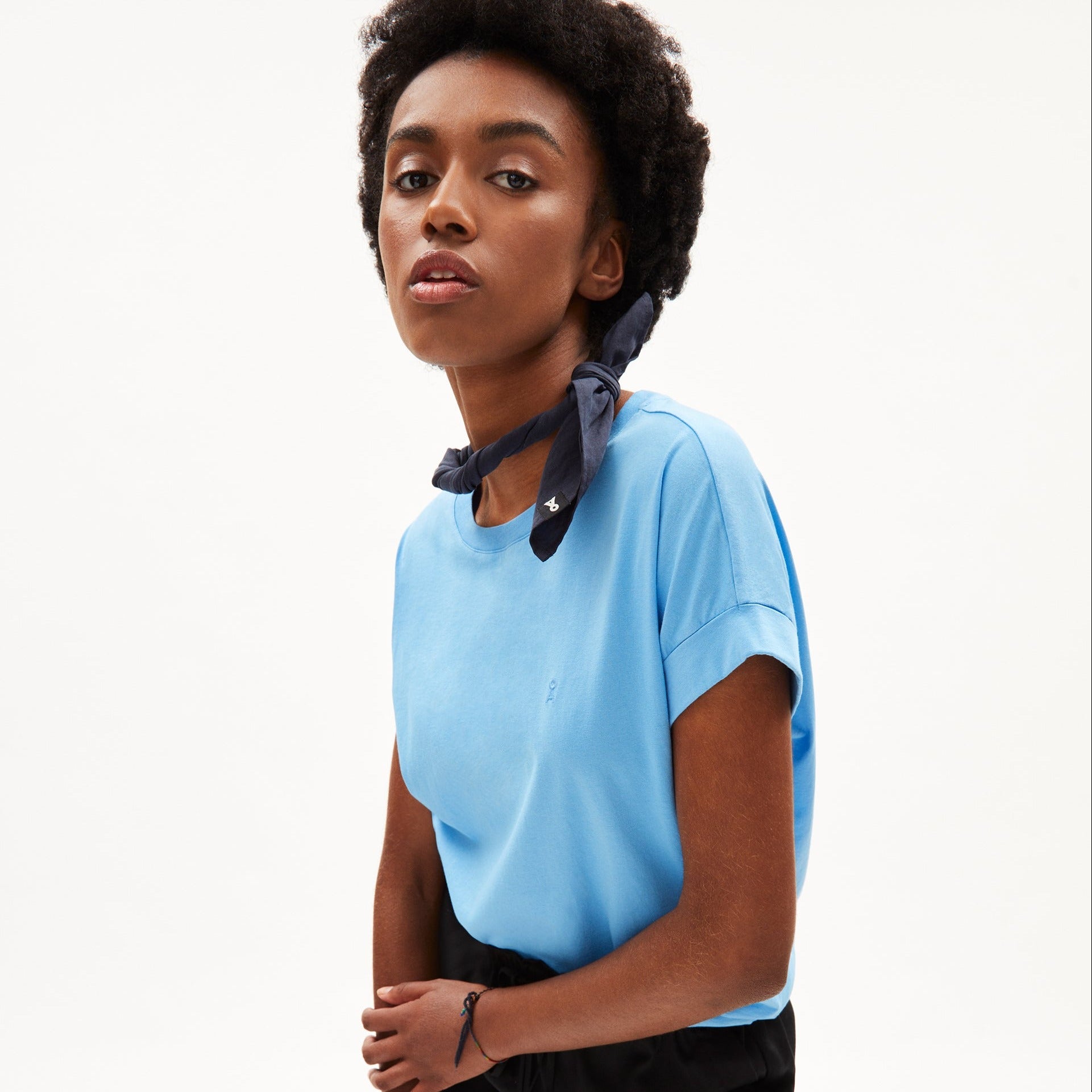 Person in blauem T-Shirt und dunklem Halstuch steht vor hellem Hintergrund.