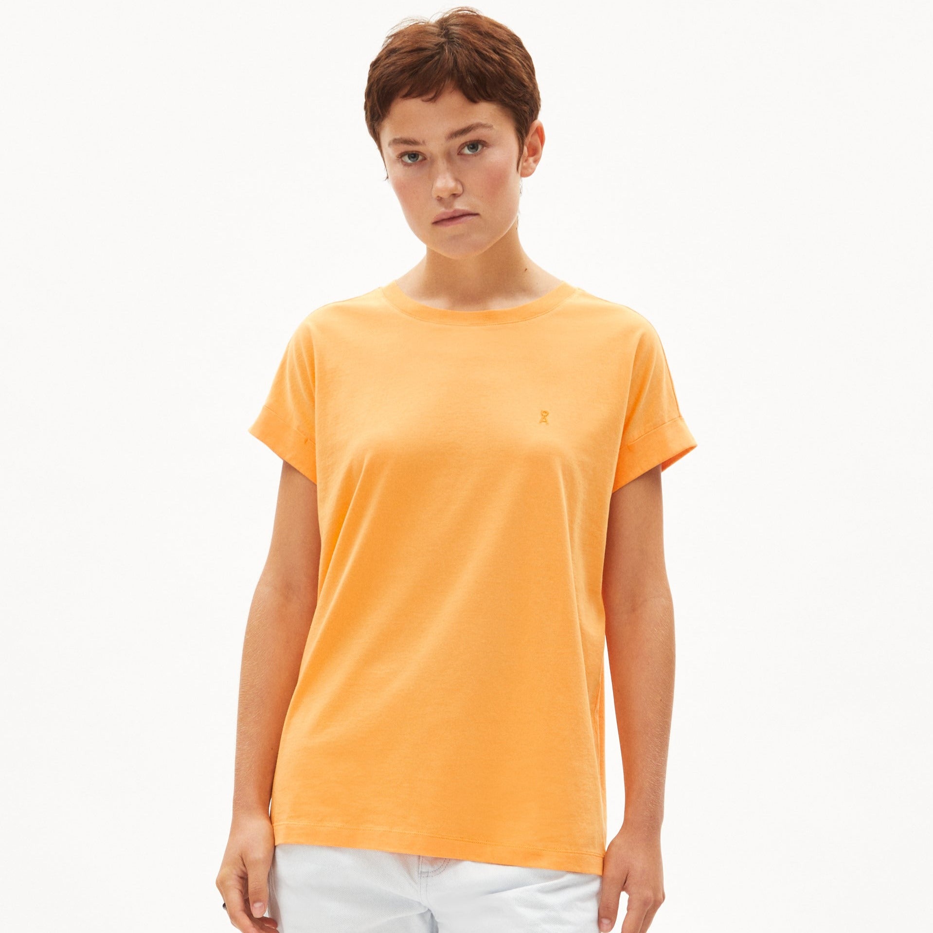 Person in an orange T-shirt and white pants stands against a plain background.