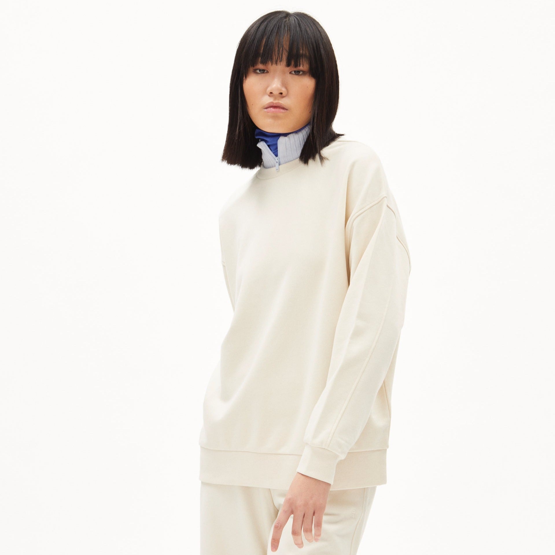 Frau in cremefarbenem Oberteil mit hochgeschlossenem blauen Kragen steht vor weißem Hintergrund.