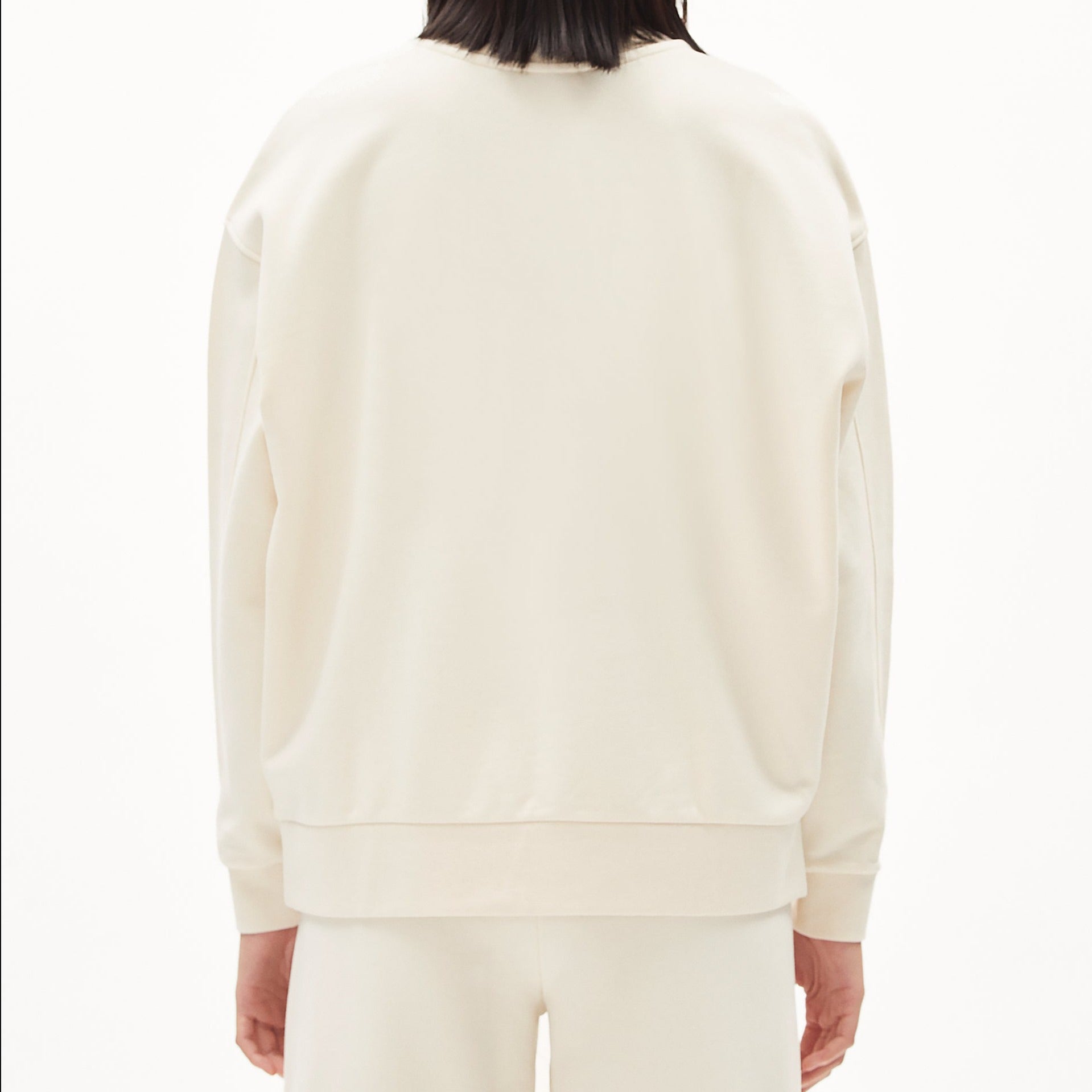 Rückansicht einer Person in einem cremefarbenen Pullover und Hose vor weißem Hintergrund.