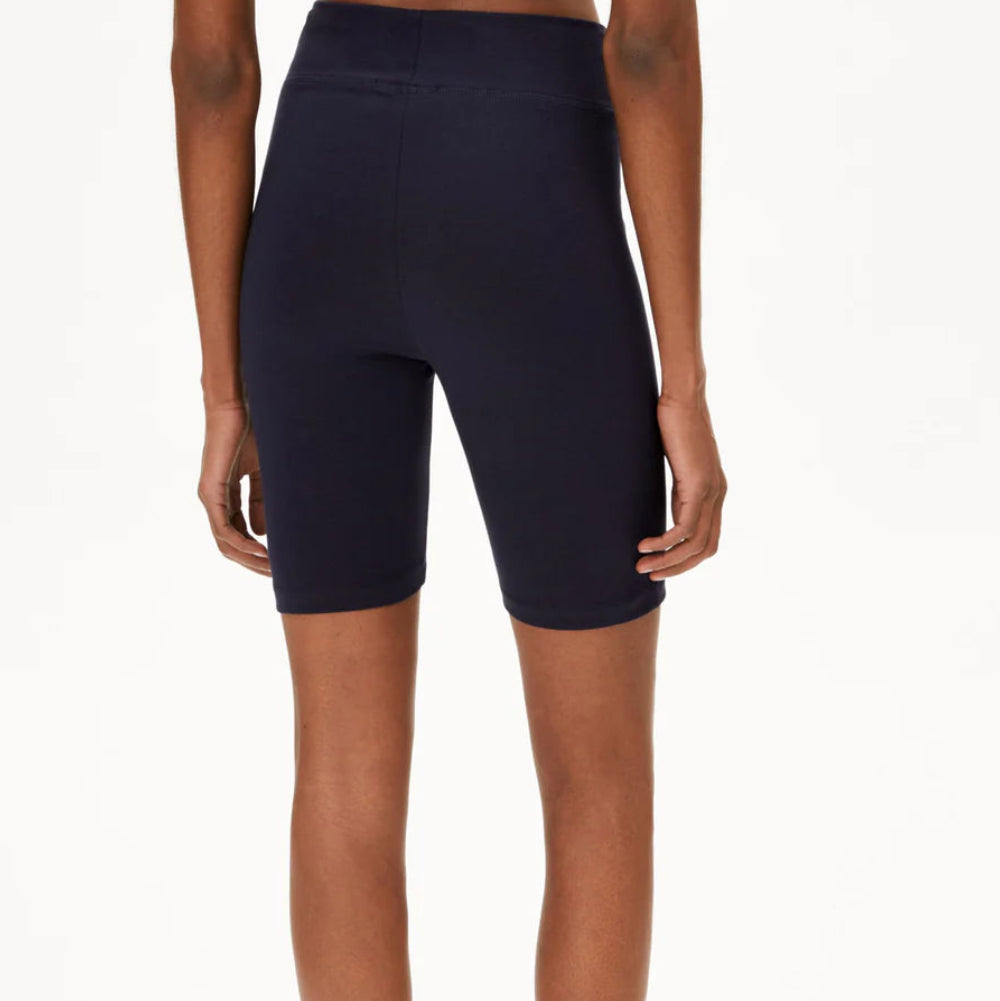 Eine Person steht mit dem Gesicht zur Wand und trägt einen weißen Sport-BH, hochgeschlossene schwarze Radlerhosen und schwarze Slide-Sandalen vor einem schlichten weißen Hintergrund.