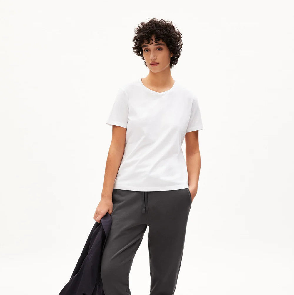 Eine Person mit kurzen lockigen Haaren trägt ein schlichtes weißes T-Shirt, eine dunkelgraue Jogginghose, weiße Socken und hellbraune Sandalen. Sie hält eine dunkle Jacke in einer Hand und steht vor einem schlichten weißen Hintergrund.