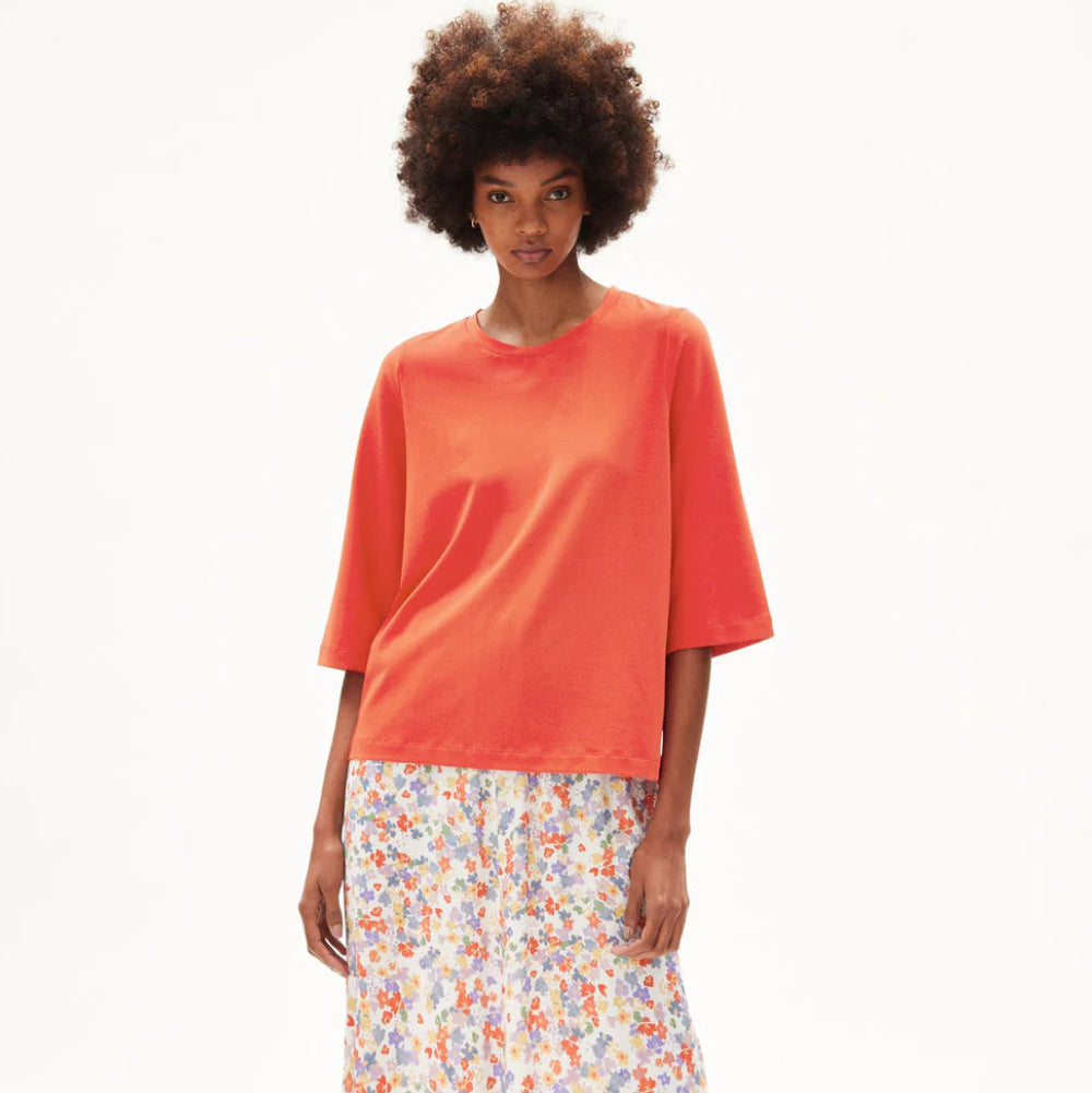 Eine Frau steht breitbeinig, trägt ein lockeres orangefarbenes kurzärmeliges Oberteil, einen weißen geblümten Midirock mit Seitenschlitz, graue Sandalen und hat lockiges Haar vor einem schlichten weißen Hintergrund.