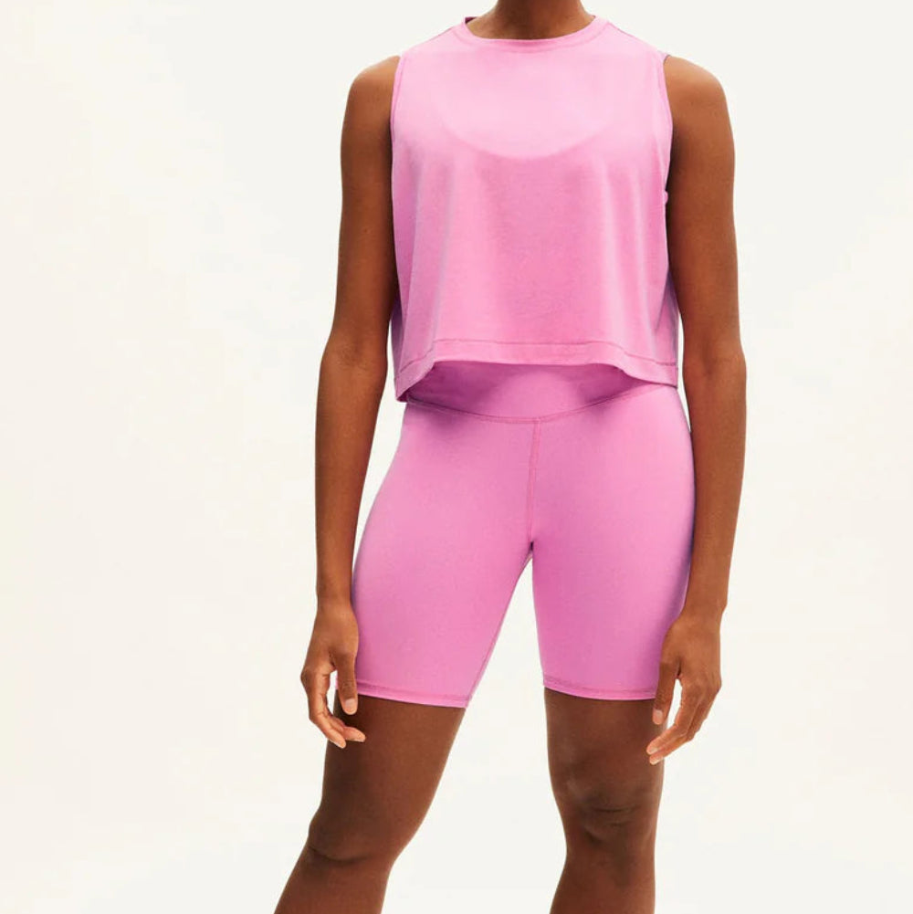 Eine Frau steht barfuß vor einem schlichten weißen Hintergrund und trägt ein lockeres, ärmelloses, hellviolettes Tank-Top über einer passenden, hoch taillierten Biker-Shorts. Sie blickt direkt in die Kamera und hat einen entspannten Gesichtsausdruck.
