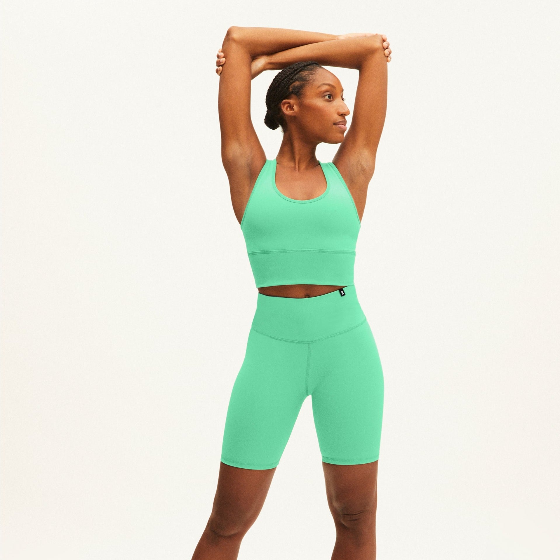 Frau in grüner Sportkleidung steht gegen einen weißen Hintergrund und streckt ihre Arme nach oben und zur Seite.