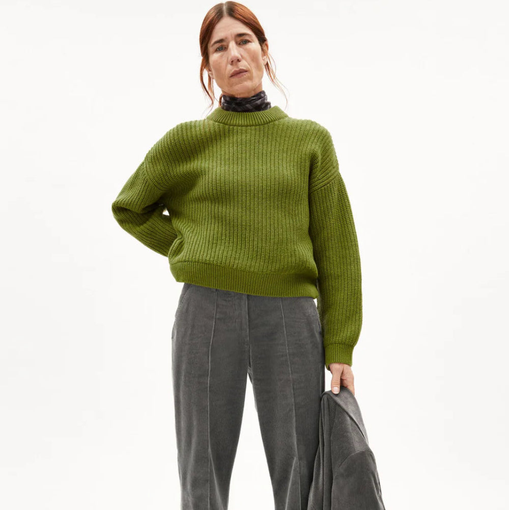 Eine Frau mit langen braunen Haaren trägt einen grünen gerippten Pullover und eine graue Hose und hält eine graue Jacke in einer Hand. Sie steht vor einem schlichten weißen Hintergrund und blickt direkt in die Kamera.