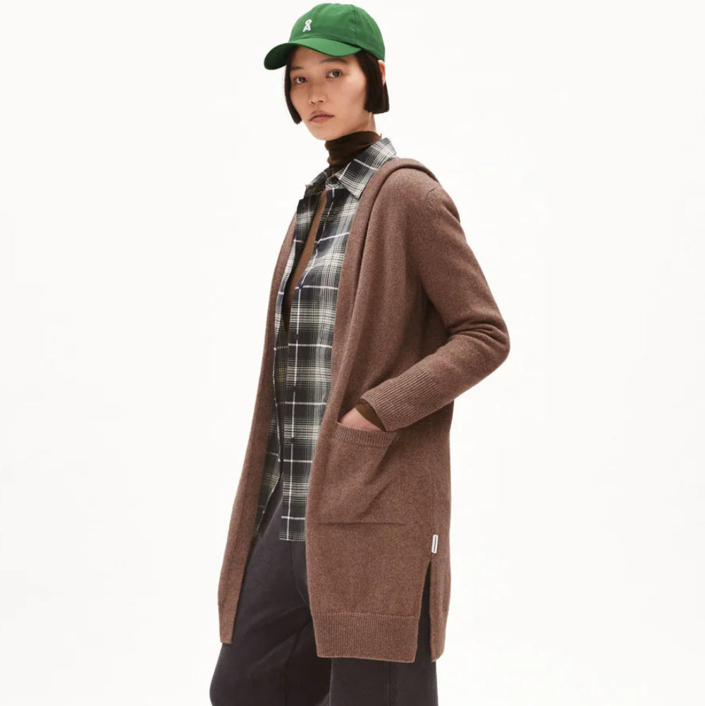 Eine Person, die eine grüne Baseballmütze, eine braune offene Strickjacke, ein kariertes Hemd, eine schwarze Hose mit weitem Bein und beigefarbene Schuhe trägt, steht mit den Händen in den Taschen der Strickjacke vor einem schlichten weißen Hintergrund.