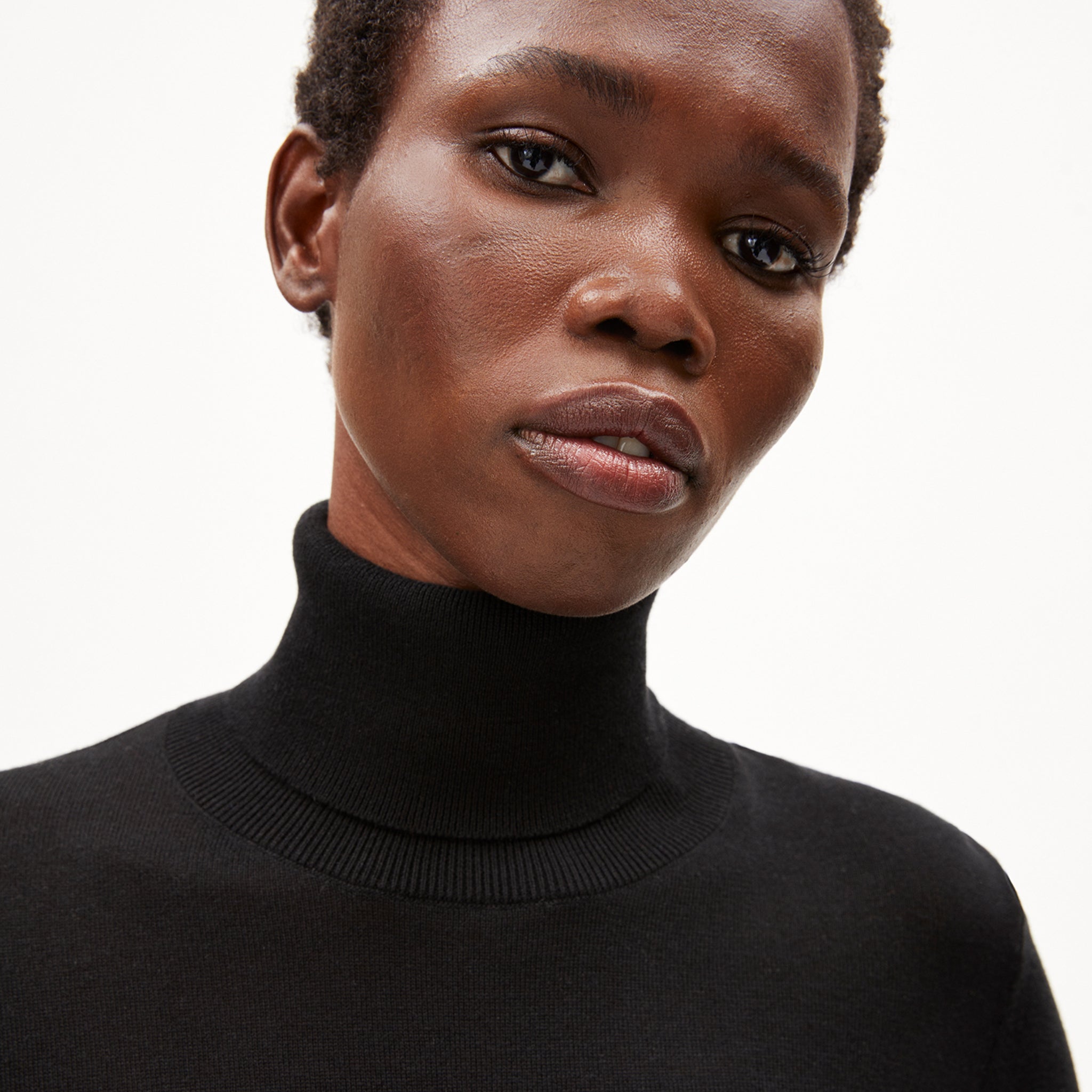 Eine Person mit kurzem, lockigem Haar trägt einen schwarzen Rollkragenpullover und schaut ruhig in die Kamera.