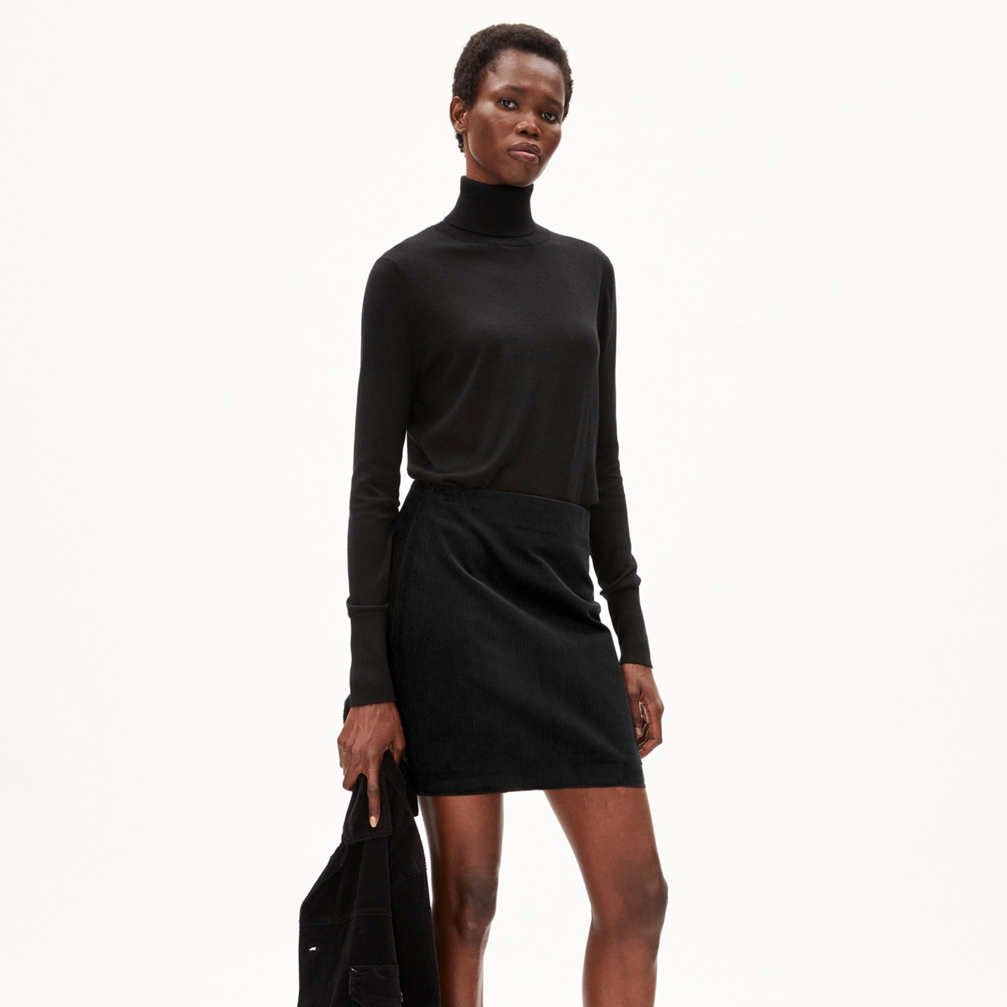 Person in schwarzem Rollkragenpullover und Rock, hält schwarze Jacke in der Hand, vor weißem Hintergrund.