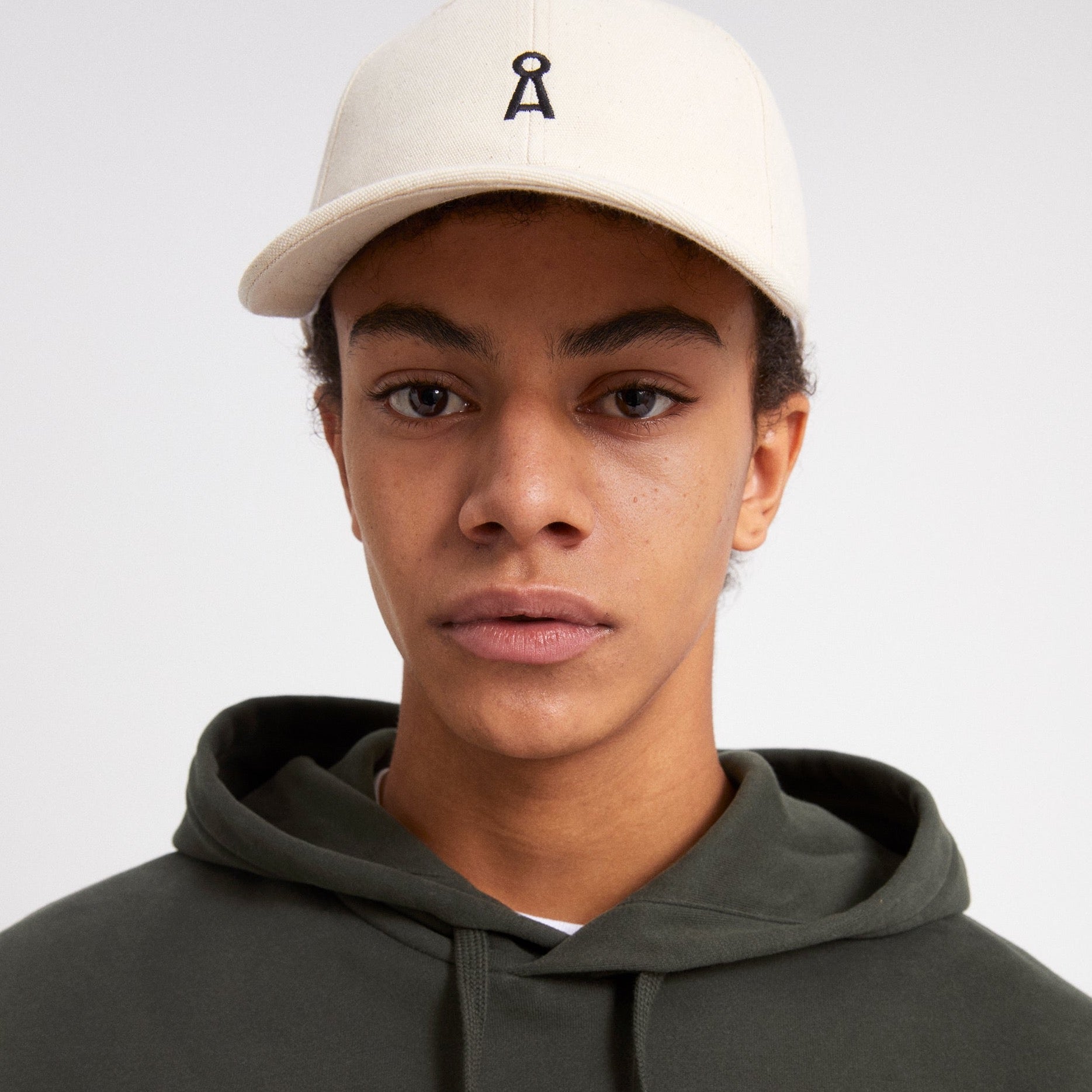 Ein Junge mit weißer Kappe und schwarzem Hoodie blickt direkt in die Kamera vor einem neutralen Hintergrund.