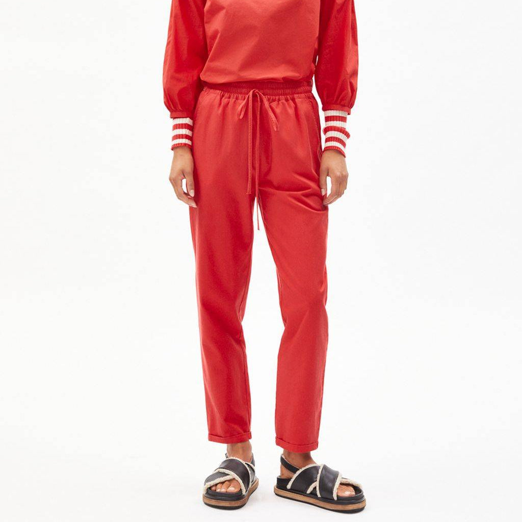 Eine Person mit kurzen, dunklen Haaren steht vor einem weißen Hintergrund und trägt ein passendes rotes, langärmeliges Oberteil und eine Hose mit Kordelzug, weiß gestreiften Bündchen und schwarzen Sandalen mit weißen Riemen.