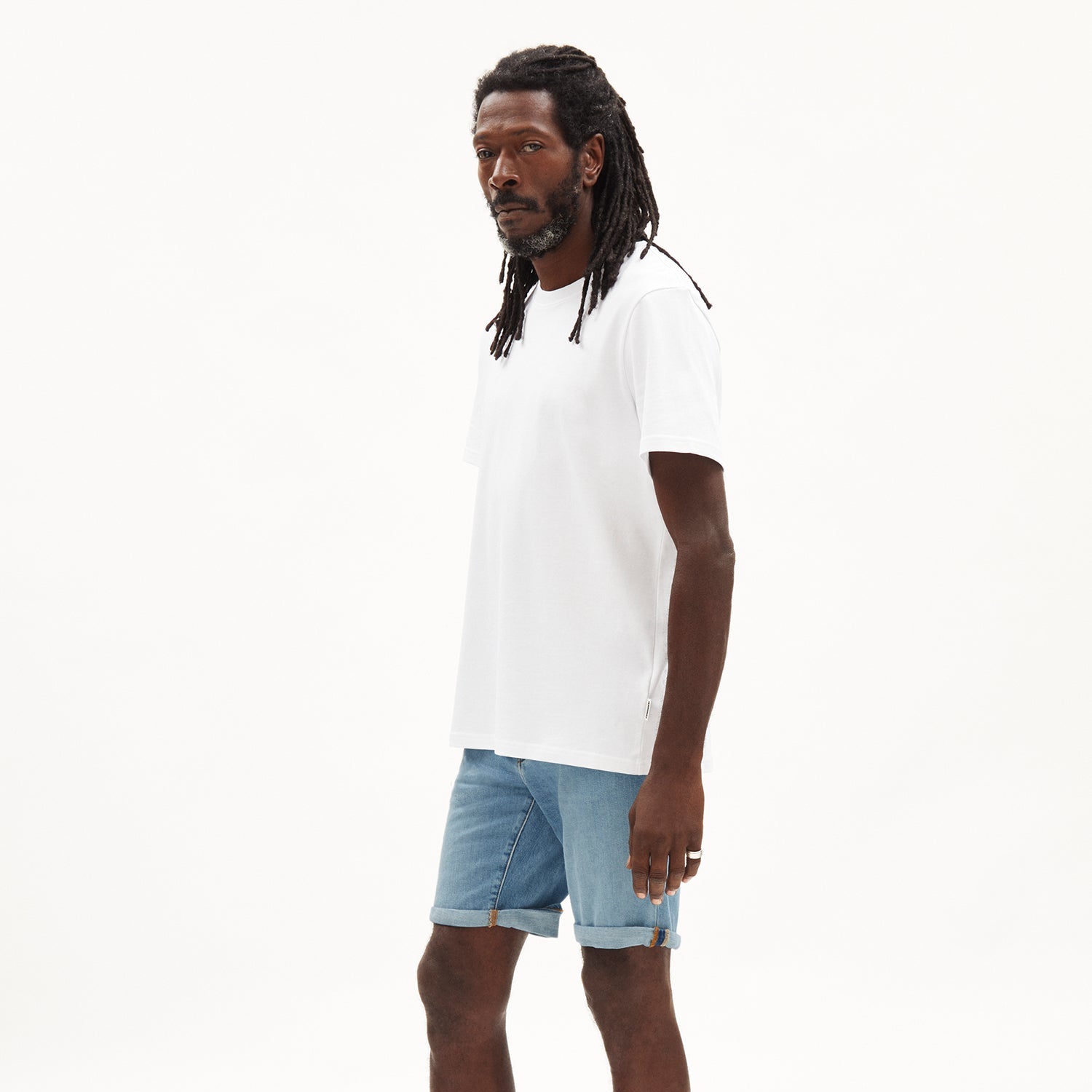 Mann mit Dreadlocks in weißem T-Shirt und Jeansshorts steht vor neutralem Hintergrund.