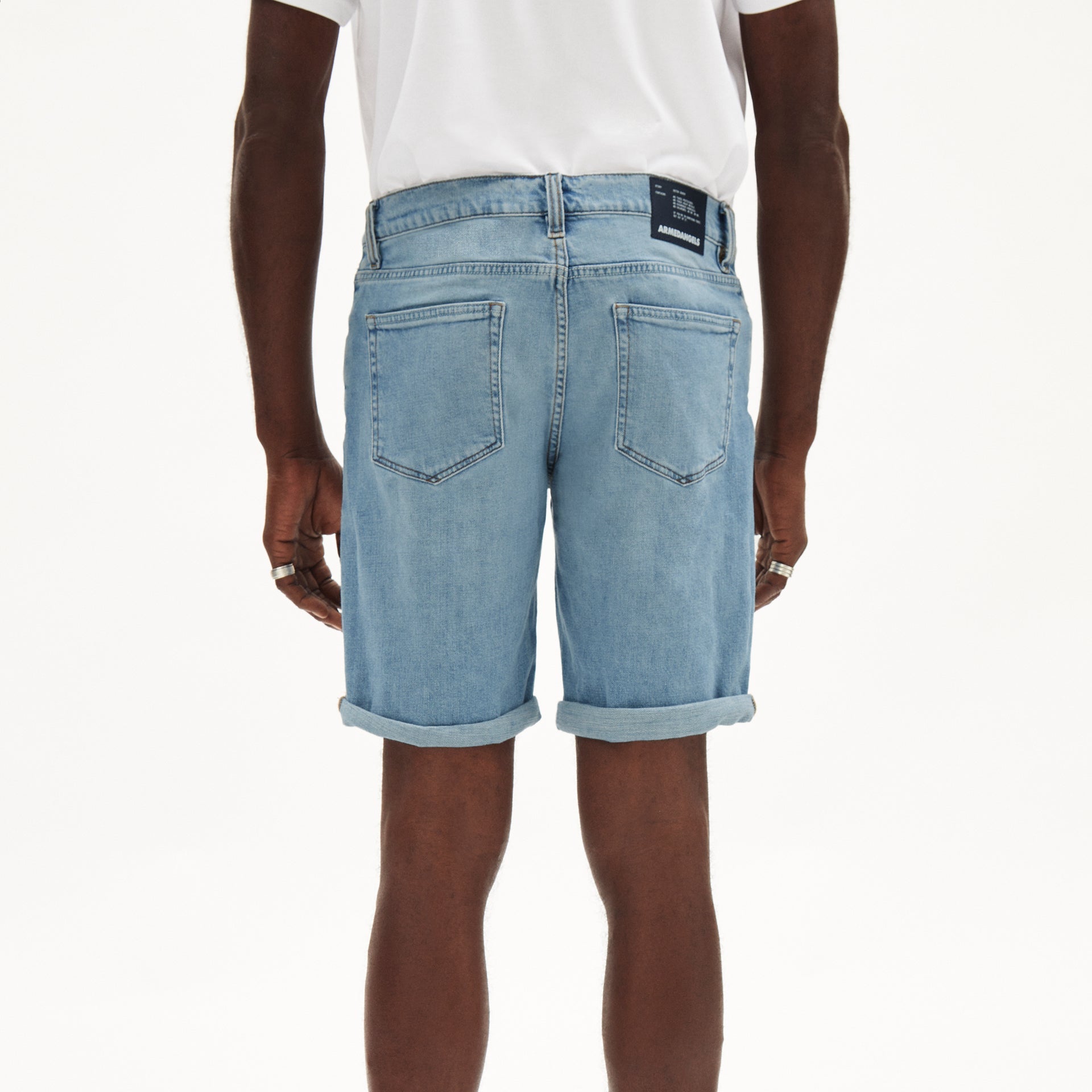 Person in weißem T-Shirt und hellblauen Jeansshorts, von hinten, mit umgeschlagenen Beinsäumen, vor hellem Hintergrund.