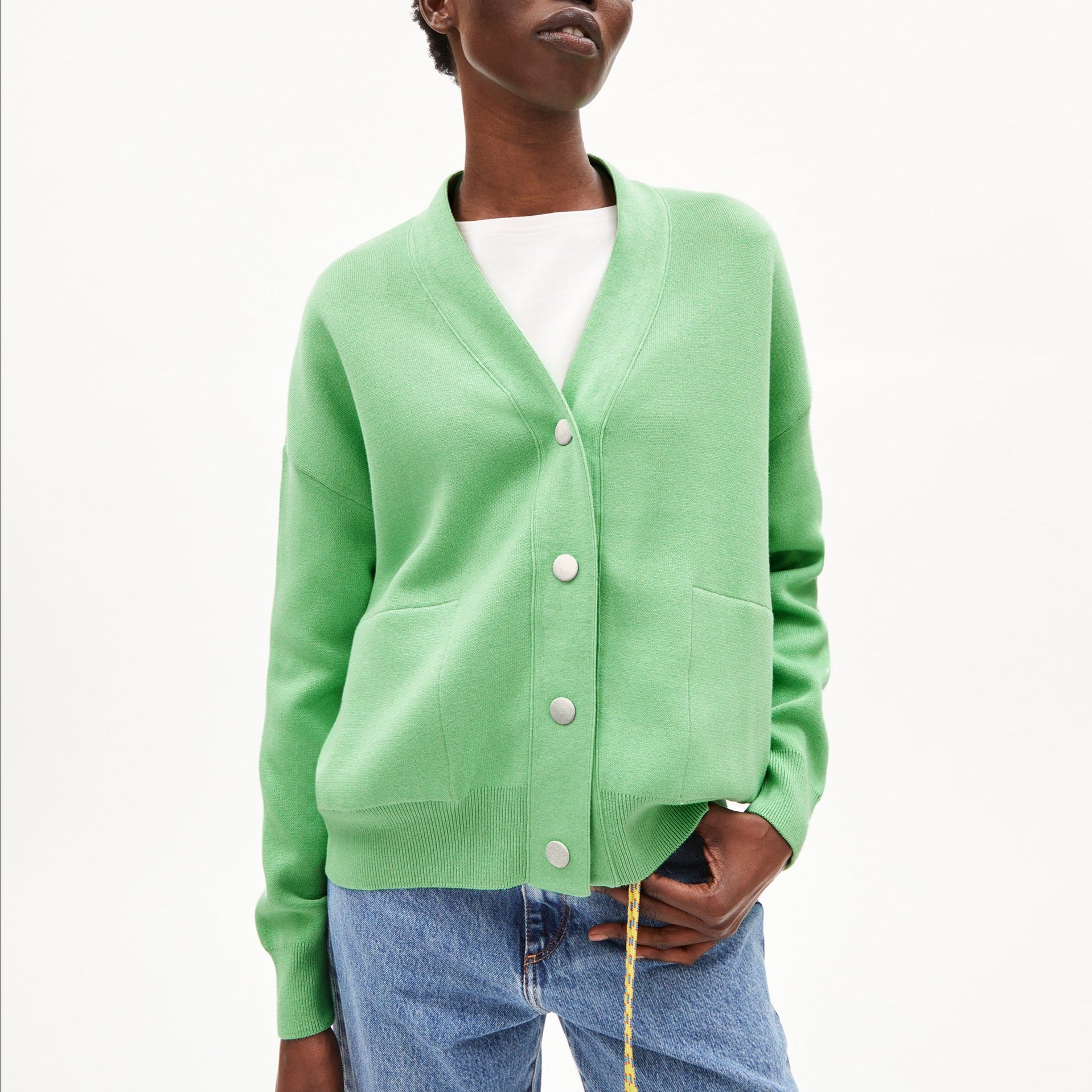Person trägt eine grüne Strickjacke mit Knöpfen, ein weißes T-Shirt und blaue Jeans vor neutralem Hintergrund.