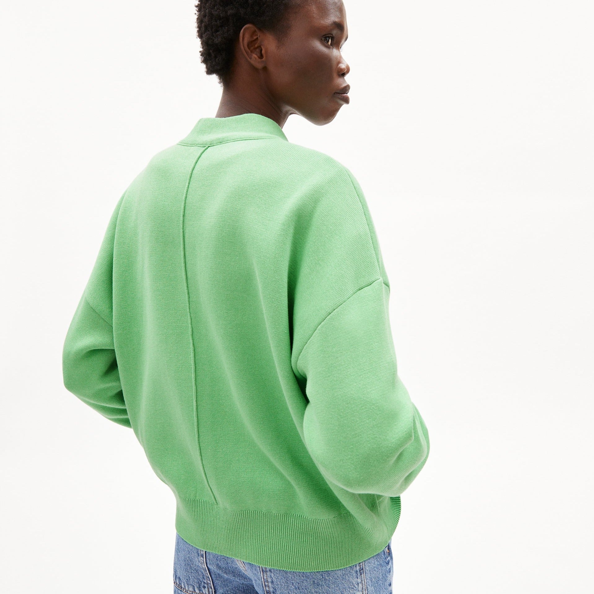 Person in grünem Pullover und blauer Jeans, von hinten fotografiert, vor weißem Hintergrund.