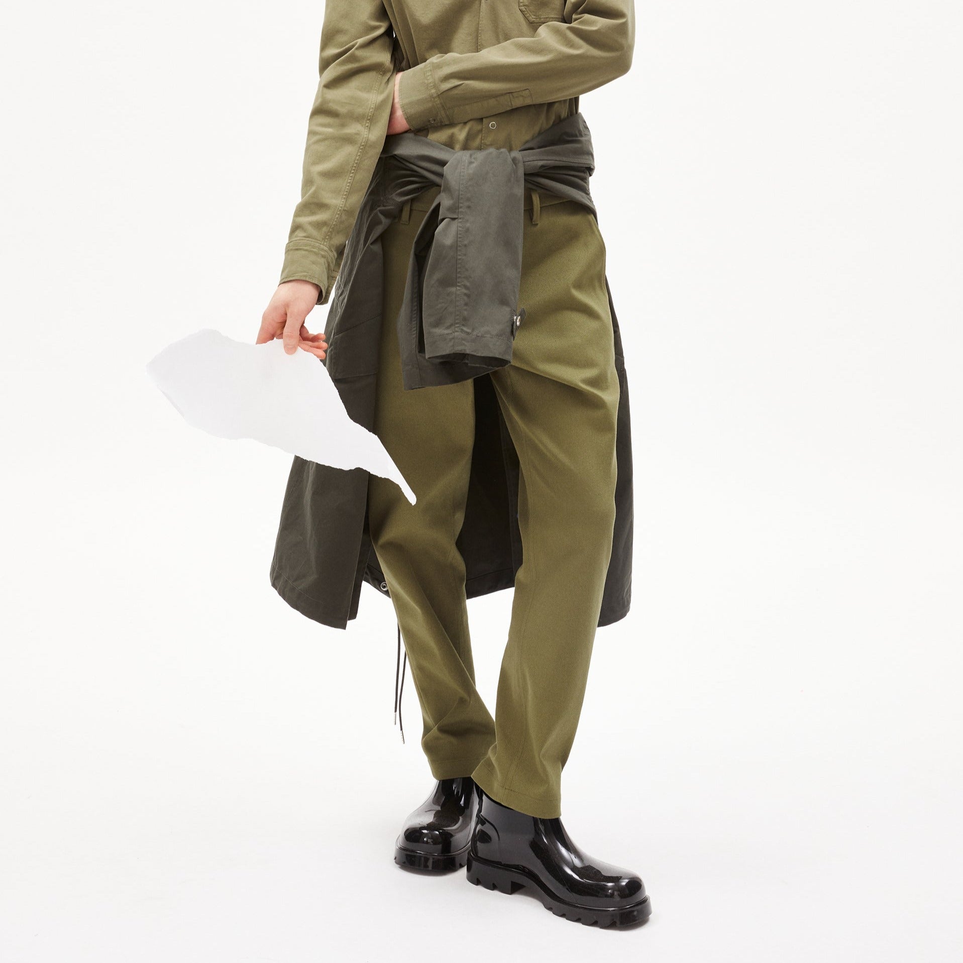 Person in grüner Kleidung hält ein Stück weißes Papier und trägt schwarze Stiefel, vor einem weißen Hintergrund.