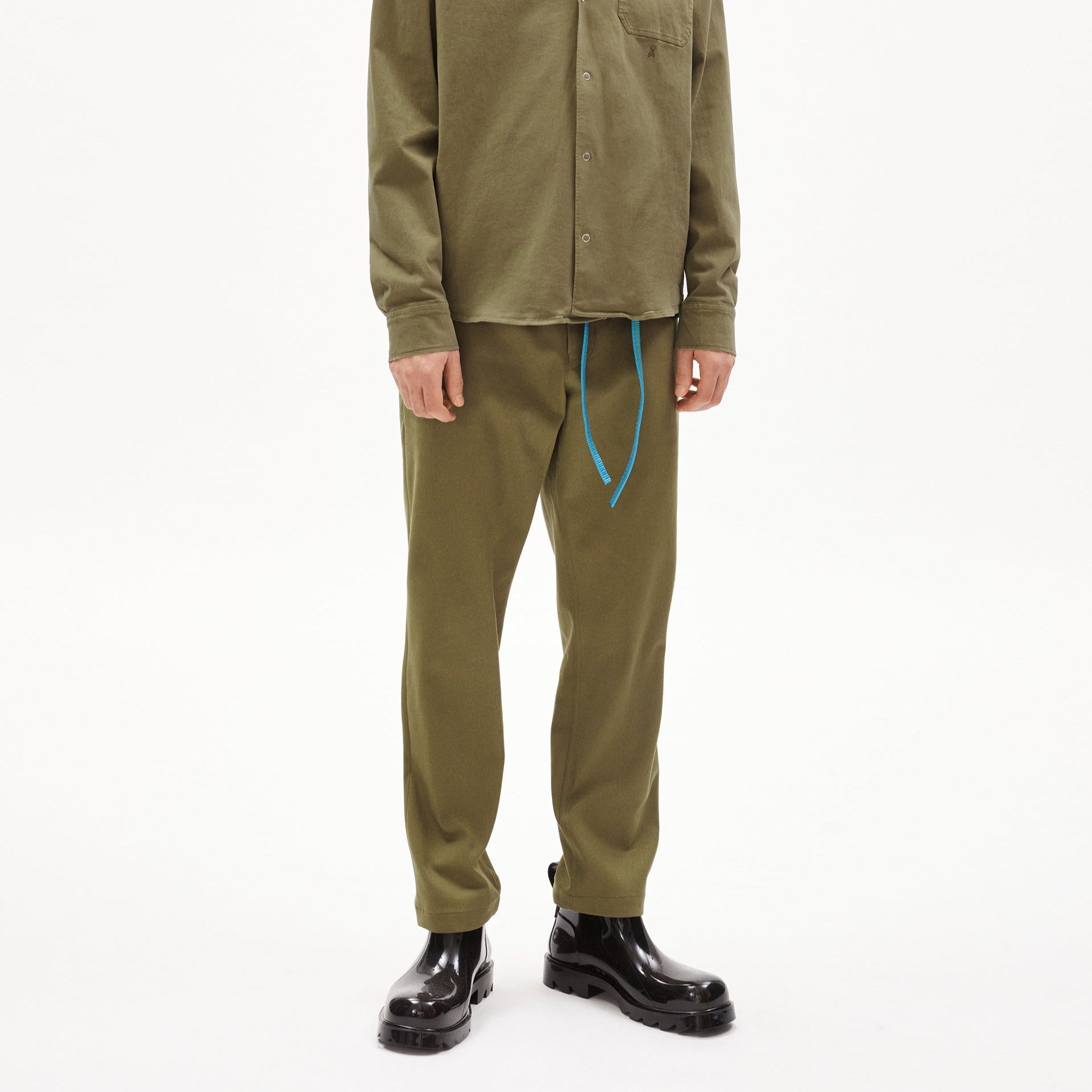 Person trägt olivgrüne Kleidung mit blauen Schnürbändern und schwarzen glänzenden Stiefeln vor weißem Hintergrund.