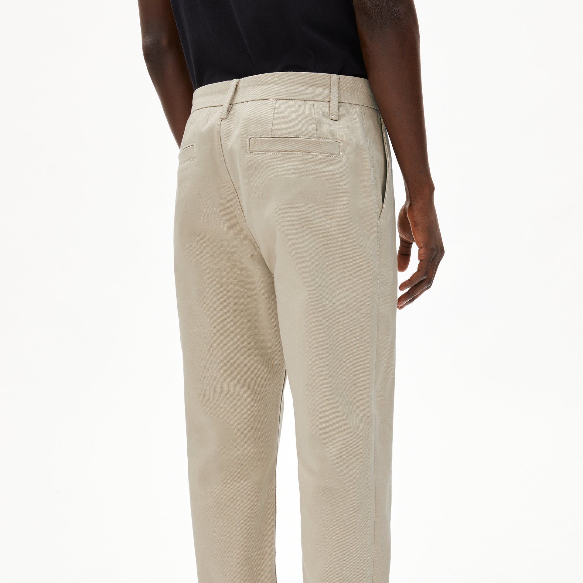 Person in beige Hosen und schwarzem Hemd von hinten fotografiert auf neutralem Hintergrund.