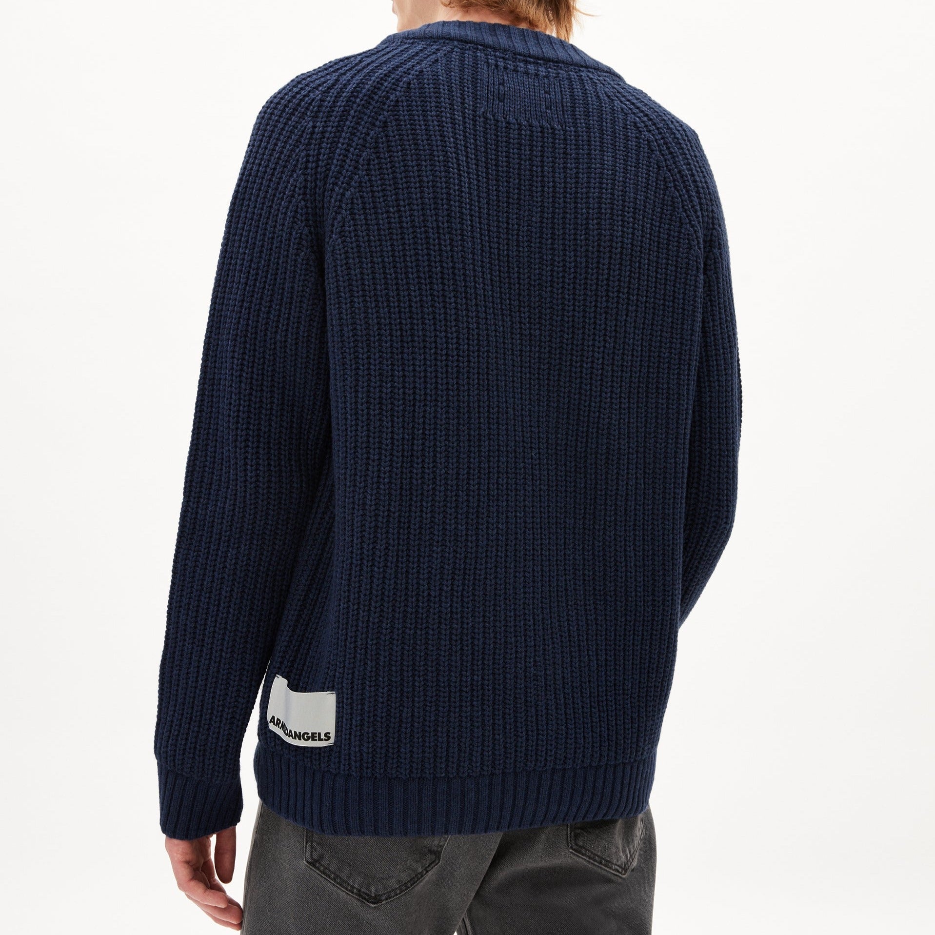 Person von hinten in dunkelblauem Strickpullover und grauen Jeans, mit einem Etikett am unteren Rücken.