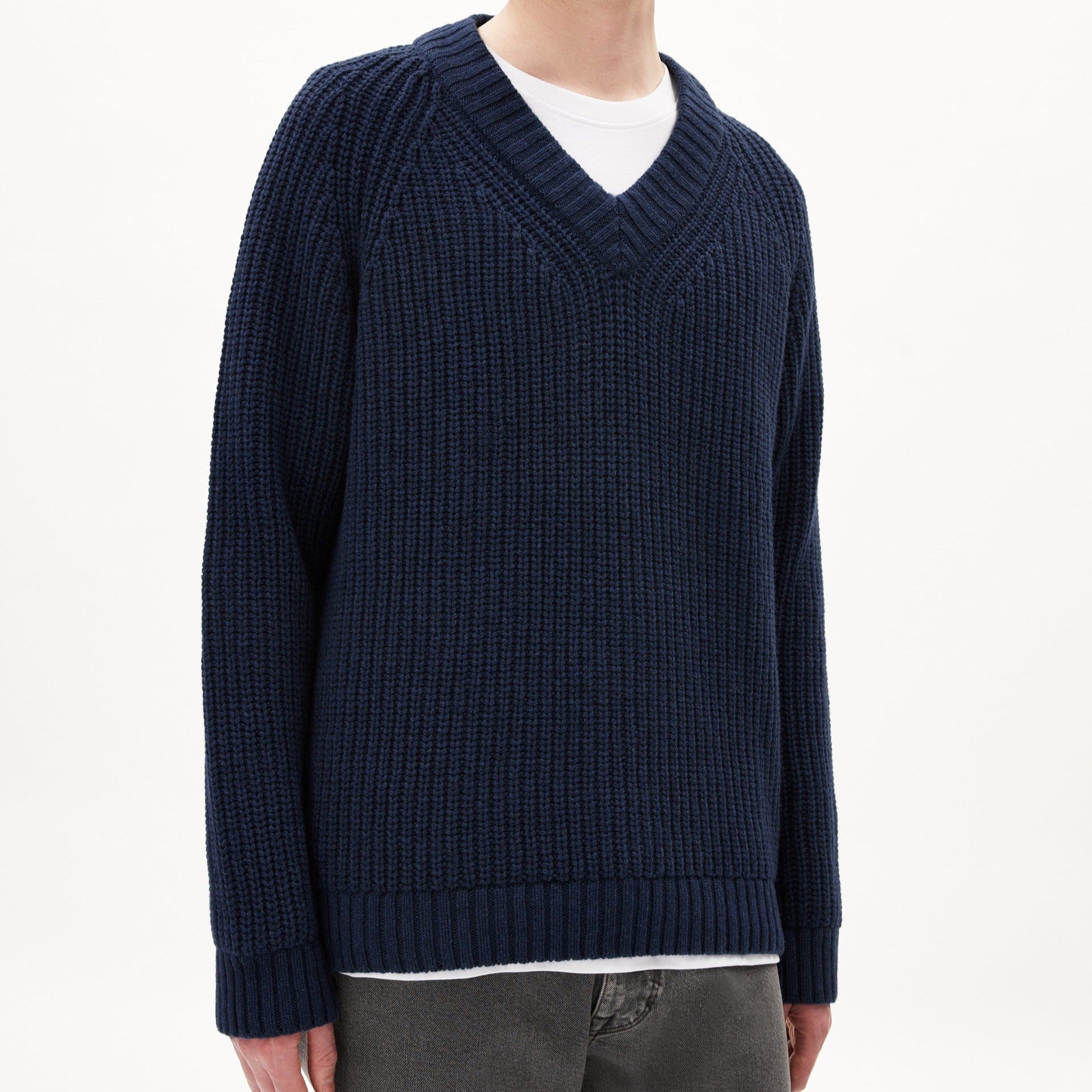 Person in a dark blue V-Pullover über einem weißen T-Shirt, teilweiser Oberkörper sichtbar, vor neutralem Hintergrund.