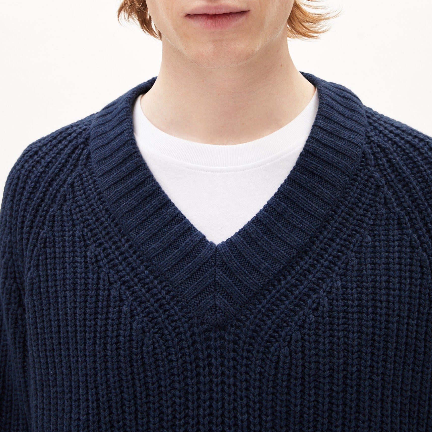 Person in dunkelblauem Strickpullover mit V-Ausschnitt und weißem T-Shirt darunter, vor hellem Hintergrund.