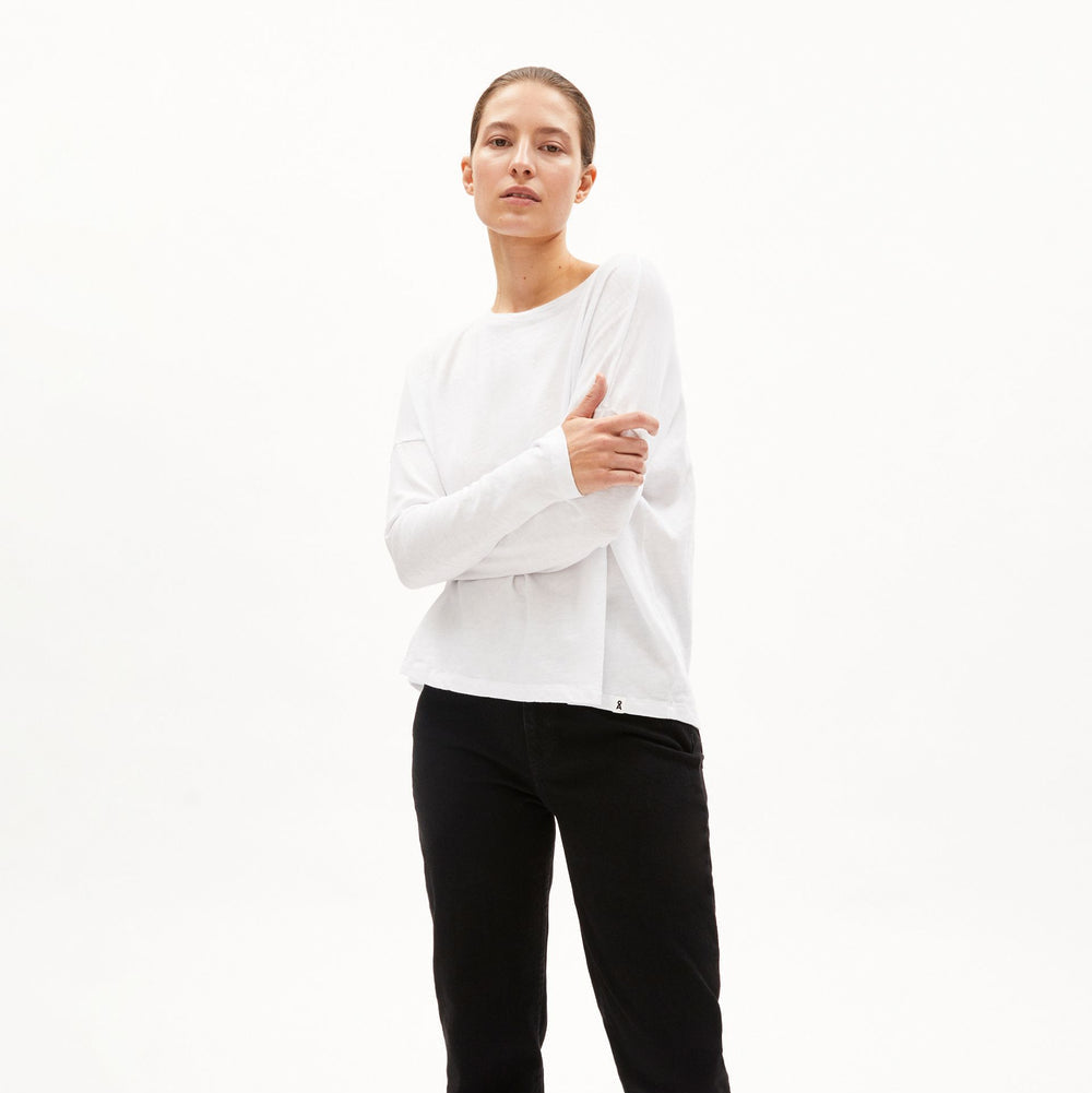 Eine Person steht vor einem schlichten weißen Hintergrund, trägt ein langärmeliges weißes Hemd, eine schwarze Hose und schwarze Clogs mit silbernen Nieten. Ihre Arme sind verschränkt und sie hat einen neutralen Ausdruck.