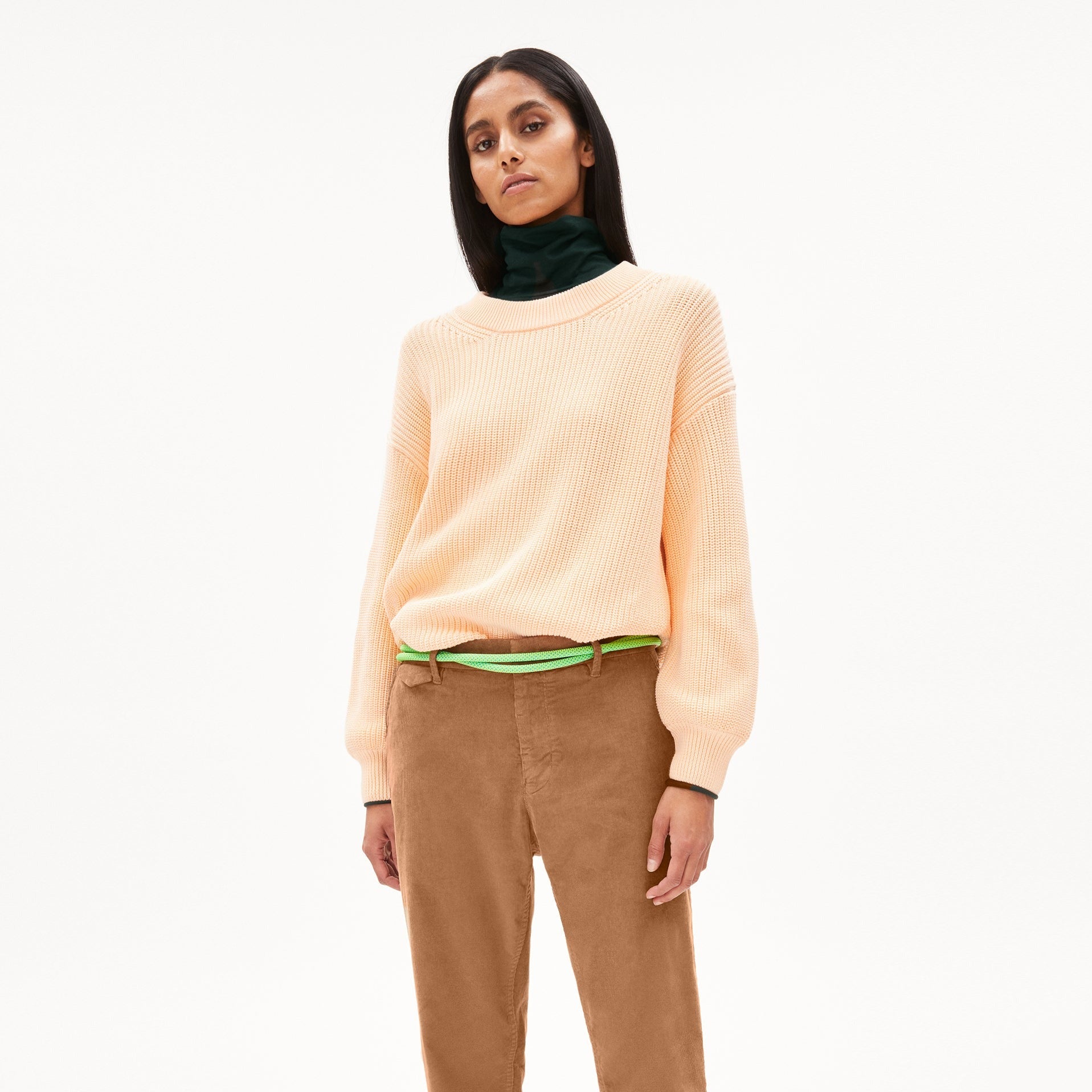 Frau in pastellfarbenem Pullover, braunen Hosen und grünem Gürtel steht vor weißem Hintergrund.