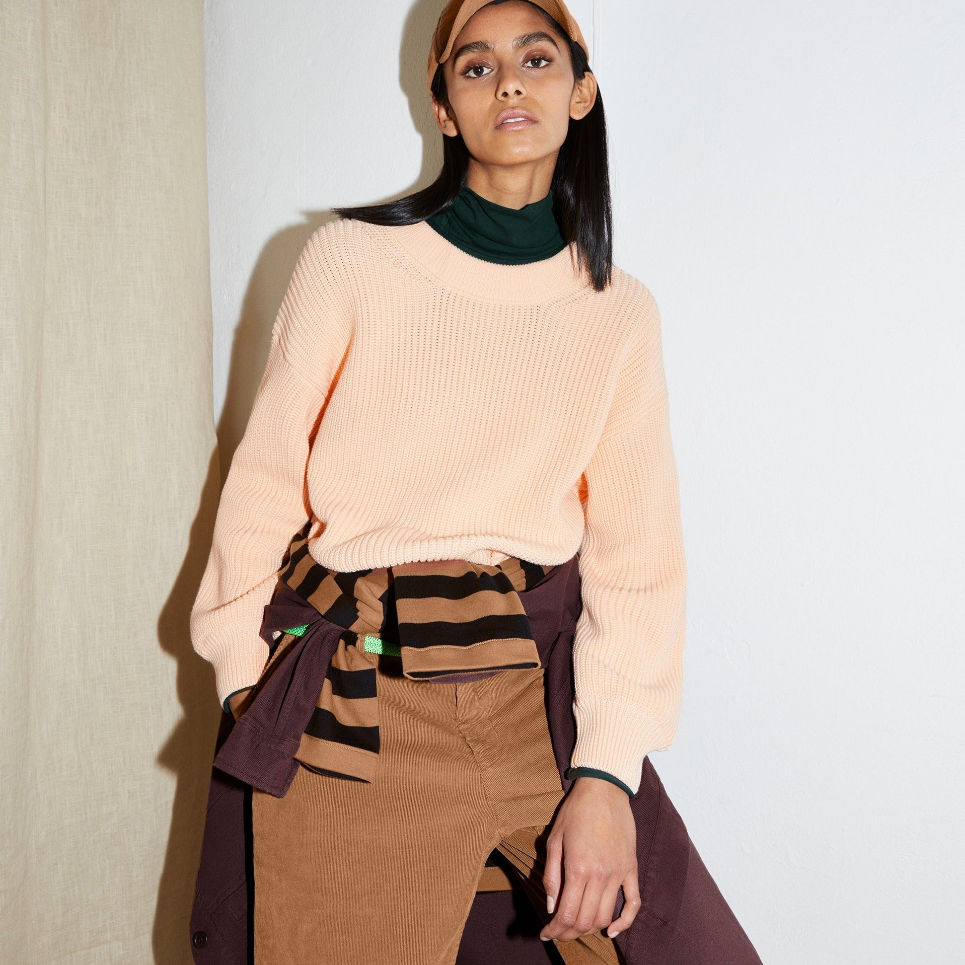 Person in peach sweater and brown pants leans against a beige wall, wearing a cap and an additional striped garment.