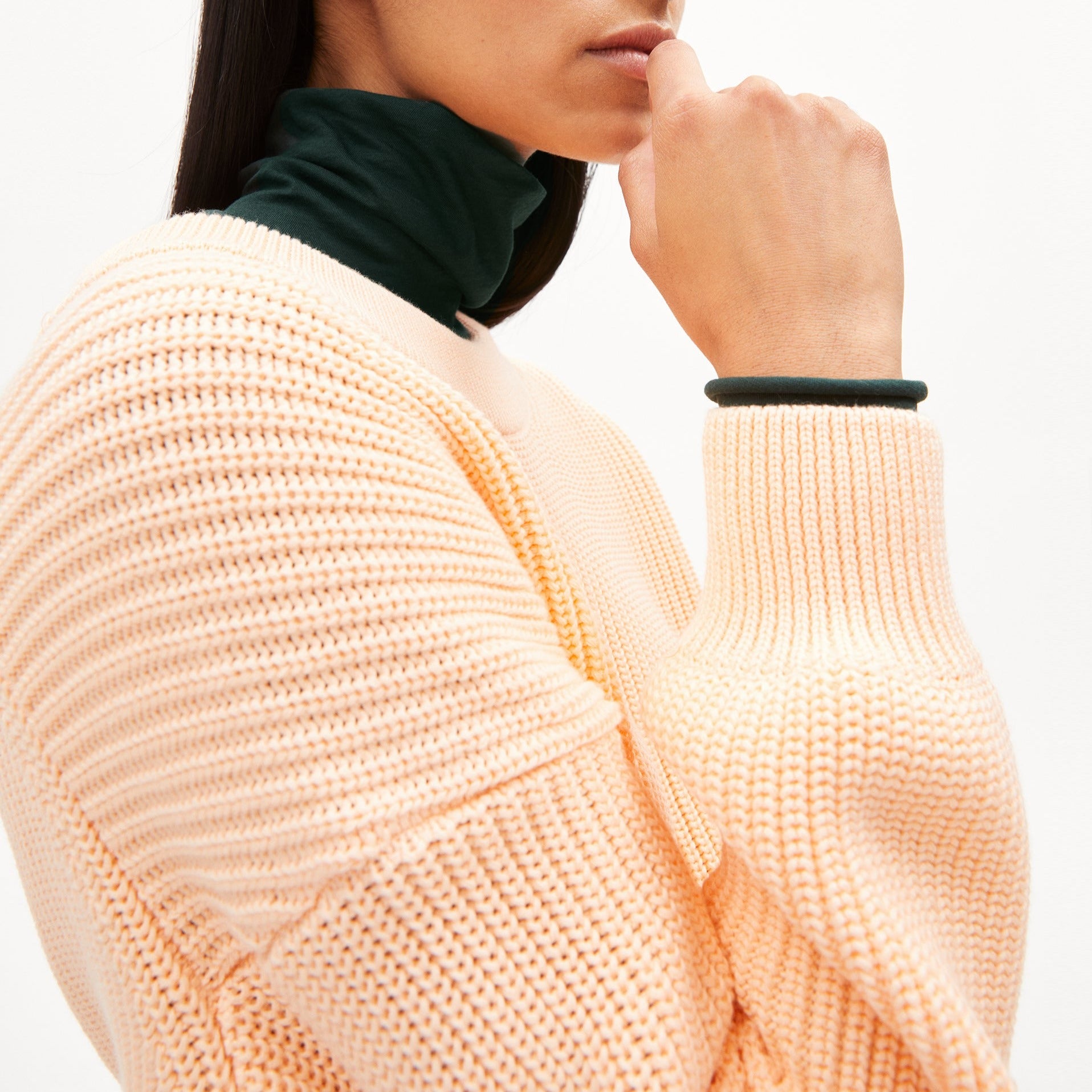 Frau in einem beigen Strickpullover, darunter ein dunkelgrüner Rollkragen, hält ihre Hand nah ans Gesicht.