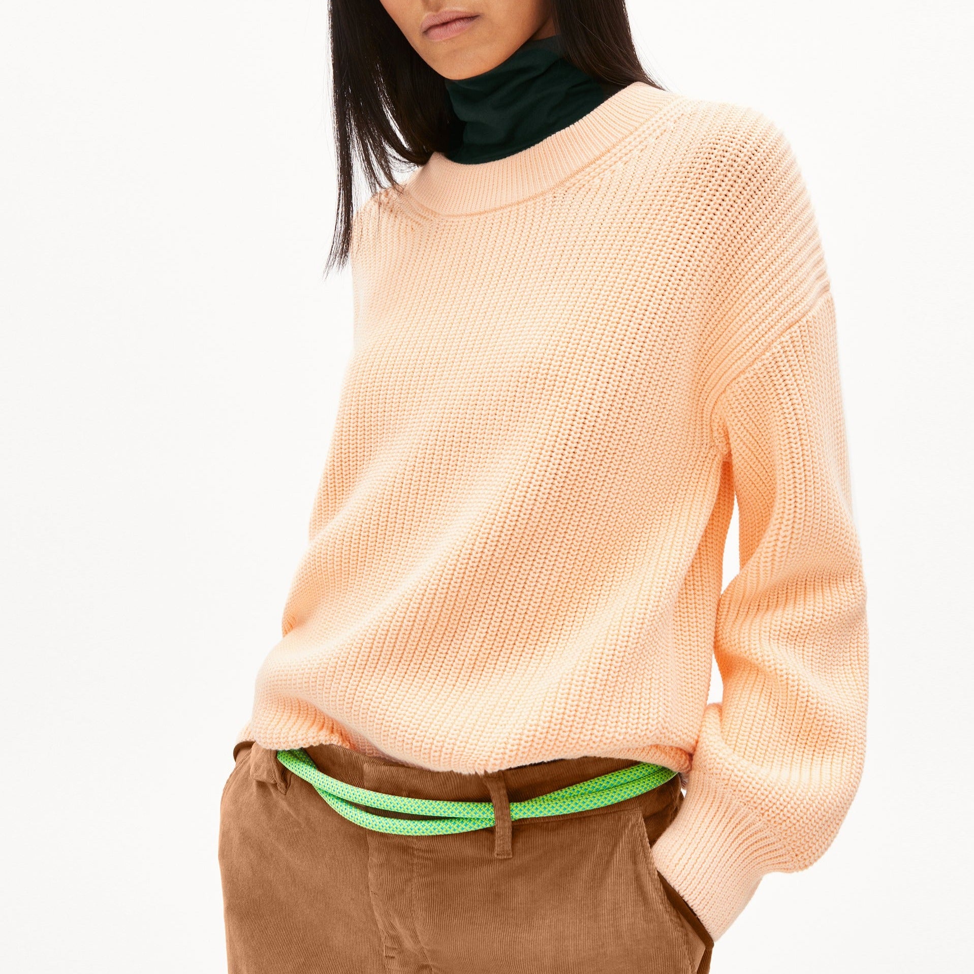 Person in einem pfirsichfarbenen Pullover und braunen Hosen mit grünem Gürtel, vor weißem Hintergrund.