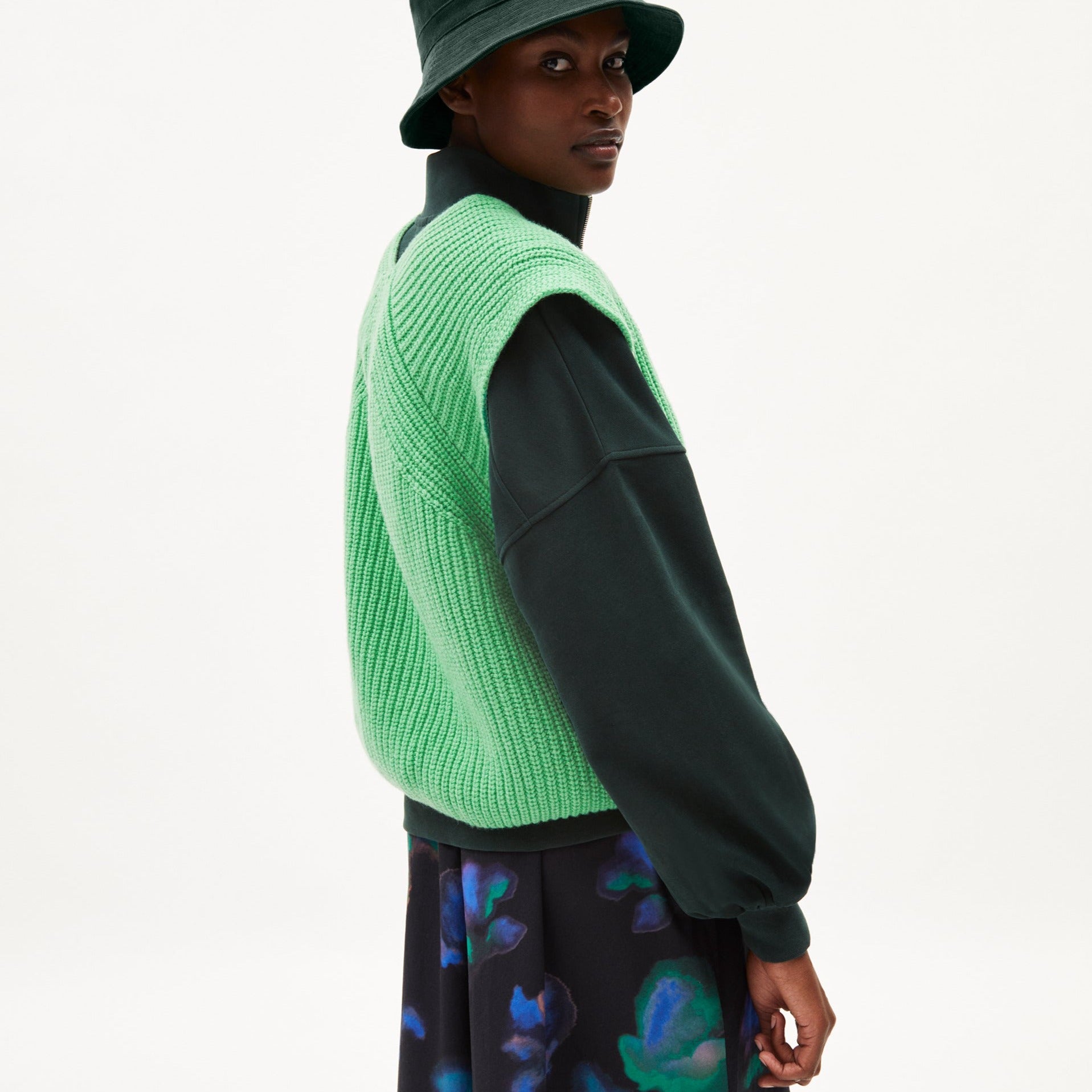 Person in einem grünen Strickpullunder, schwarzem Oberteil und Hut, schaut über die Schulter in die Kamera.