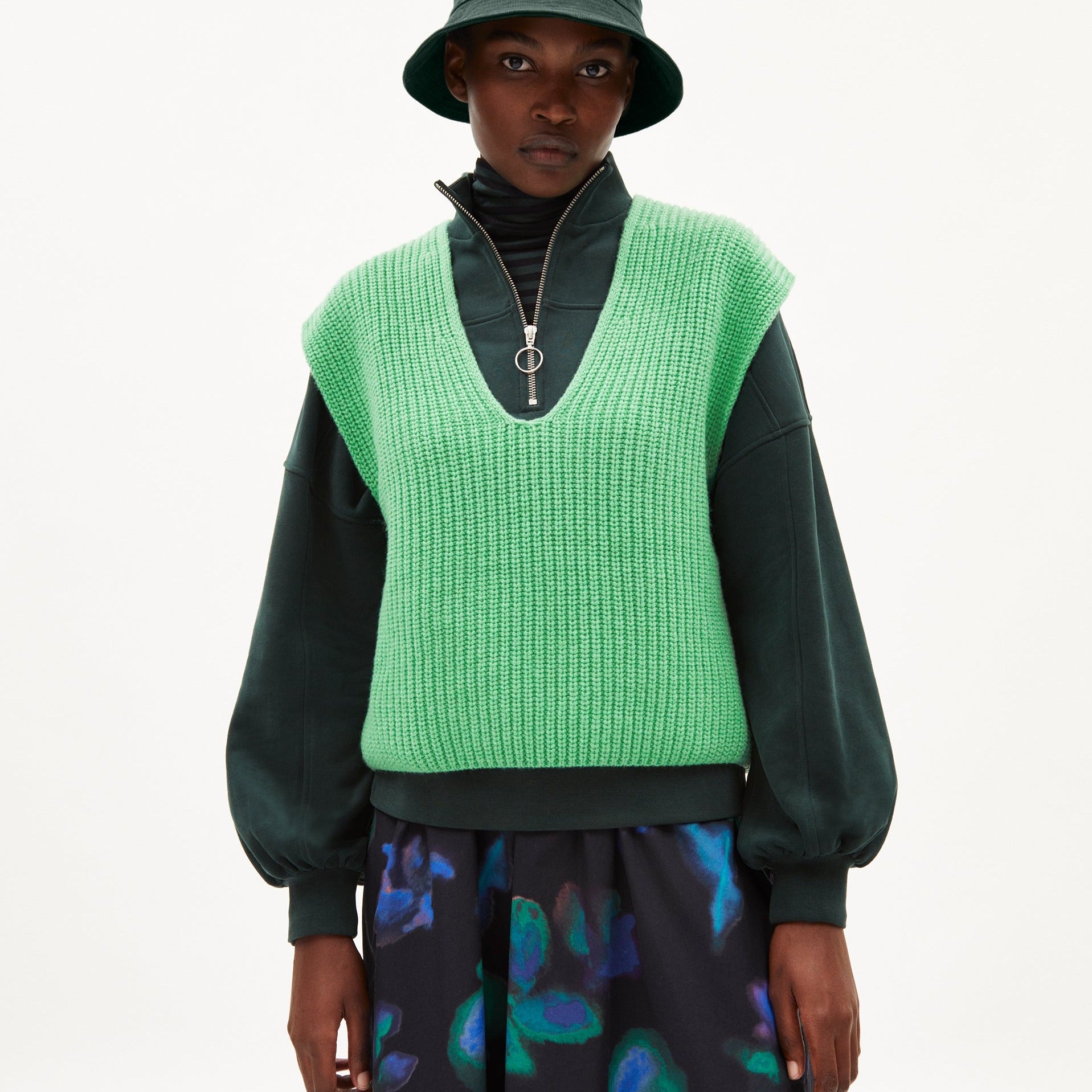 Person in grünem Strickwesten und Fischerhut, trägt schwarzen Pullover und Rock mit buntem Muster, neutraler Hintergrund.