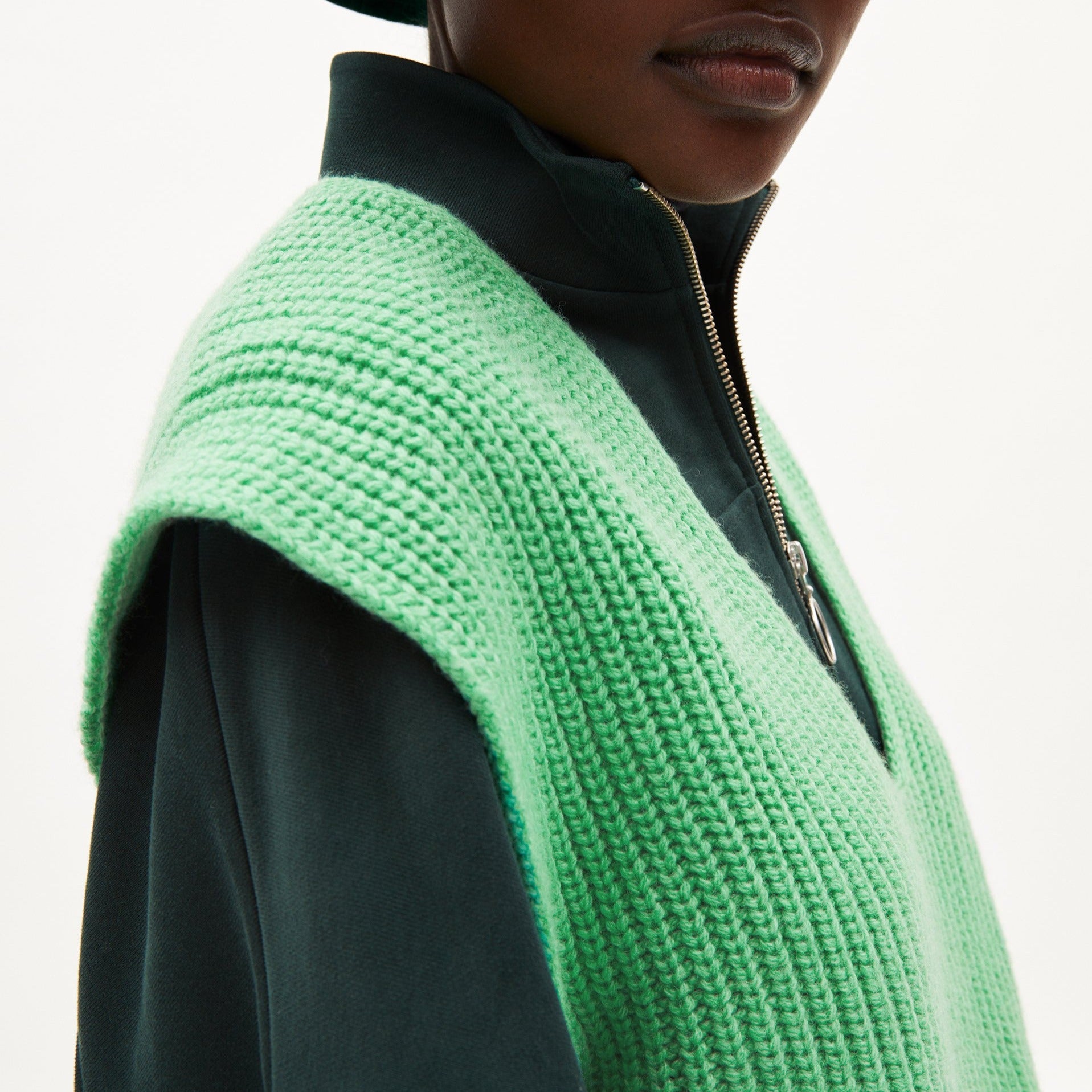 Person trägt eine grüne Strickweste über einem dunkelgrünen Oberteil mit Reißverschluss.
