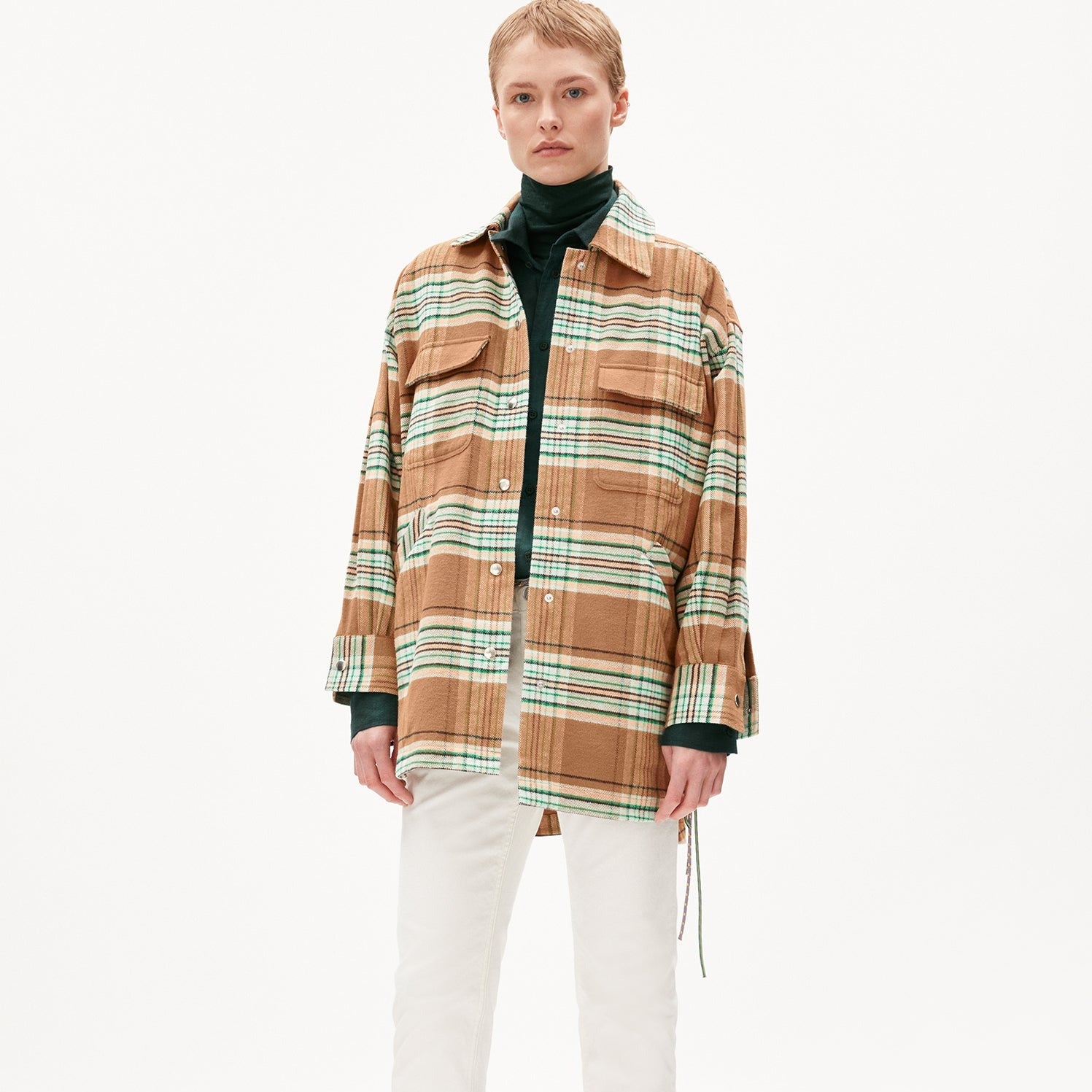 Person in kariertem Overshirt, dunklem Rollkragenpullover und weißer Hose vor weißem Hintergrund.