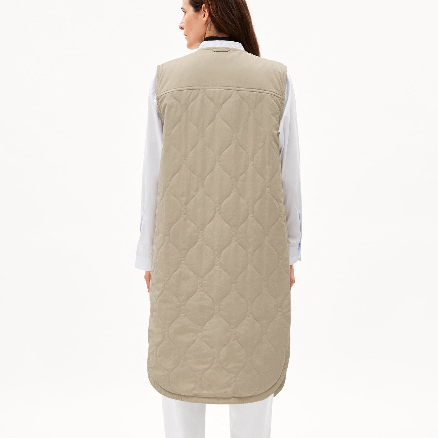 Frau im langen, beige gesteppten ärmellosen Mantel von hinten, darunter ein weißes Hemd.