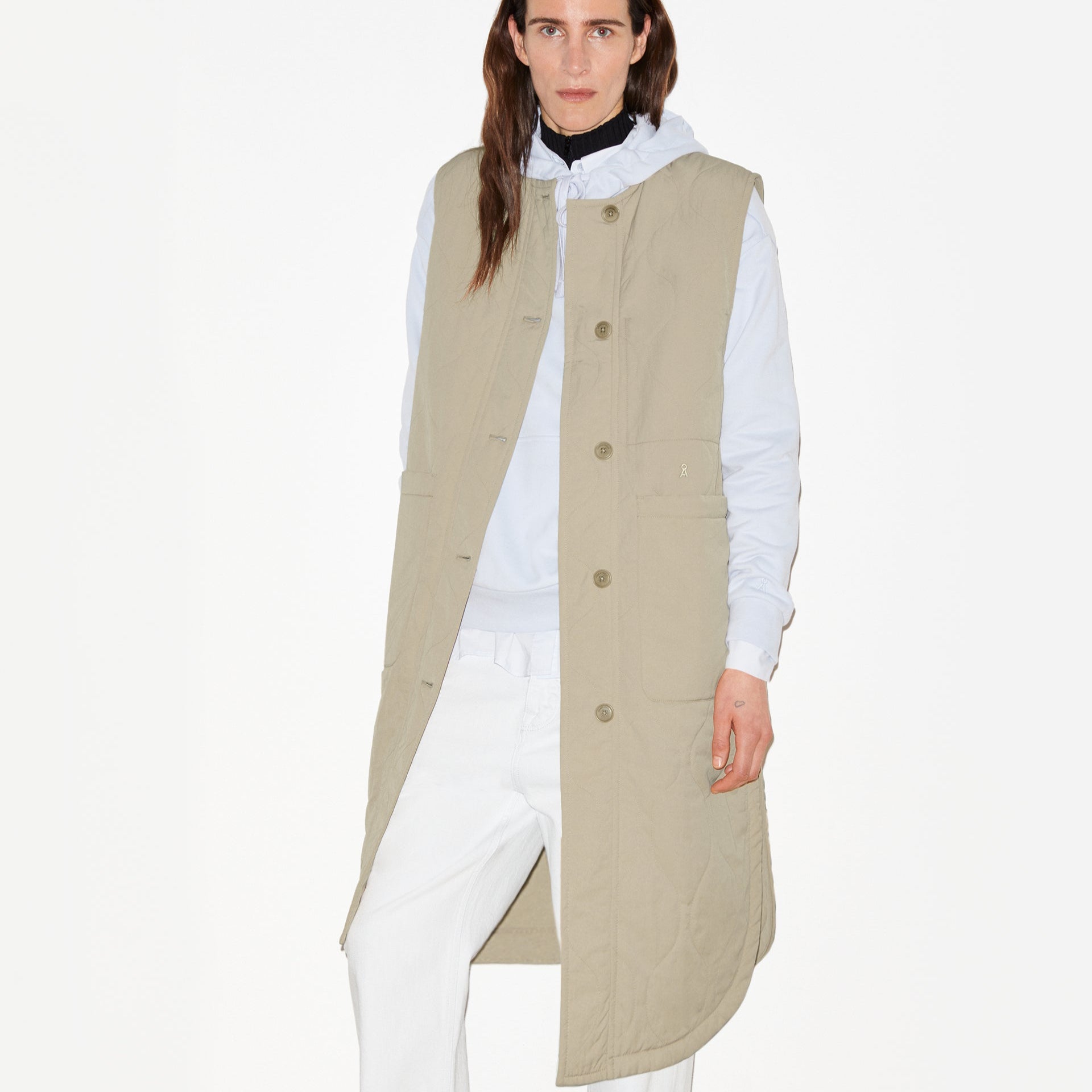Person in beige, ärmelloser Jacke und weißem Hoodie vor weißem Hintergrund.