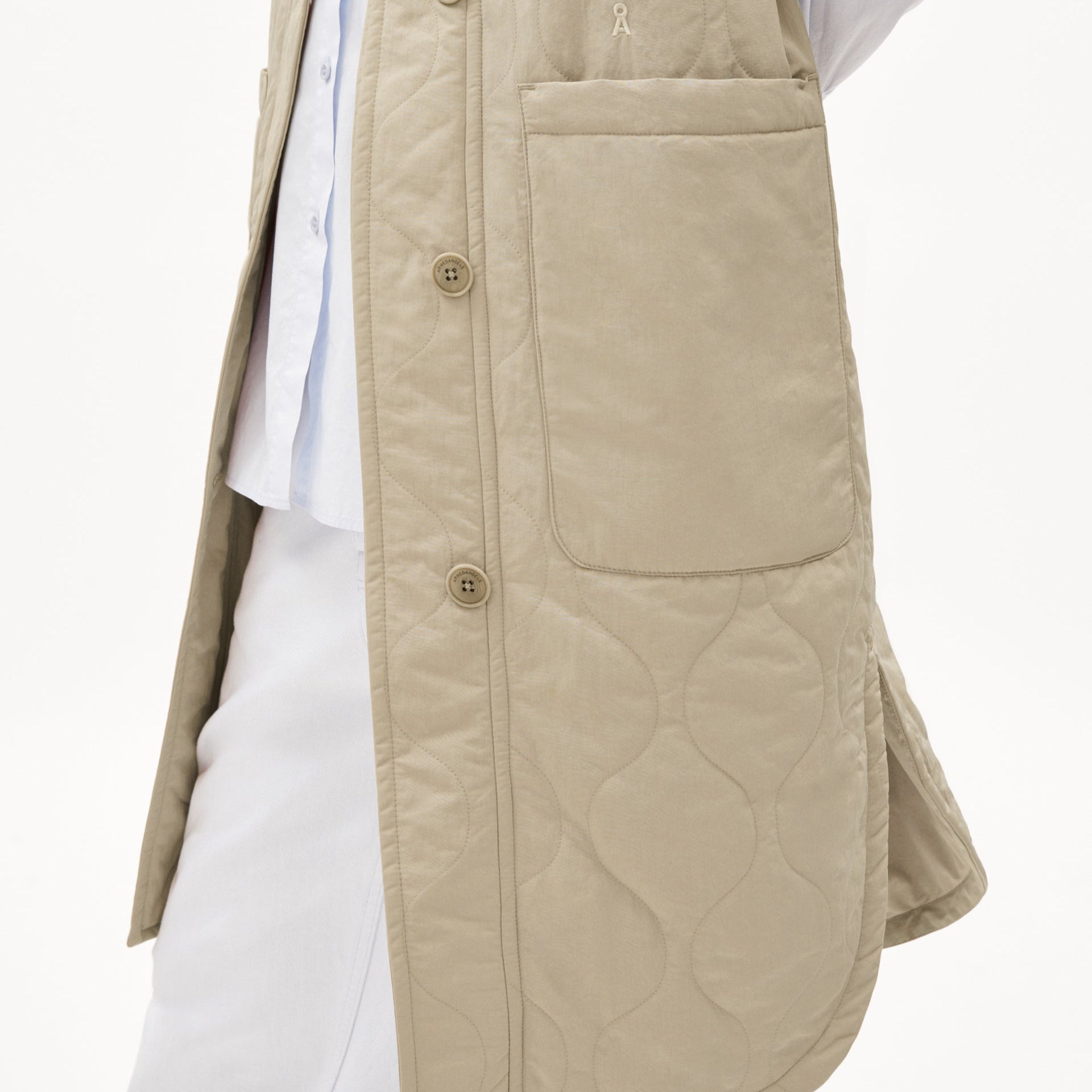 Beiger, gesteppter Mantel mit großen Taschen und Knöpfen über einem weißen Outfit vor einem weißen Hintergrund.