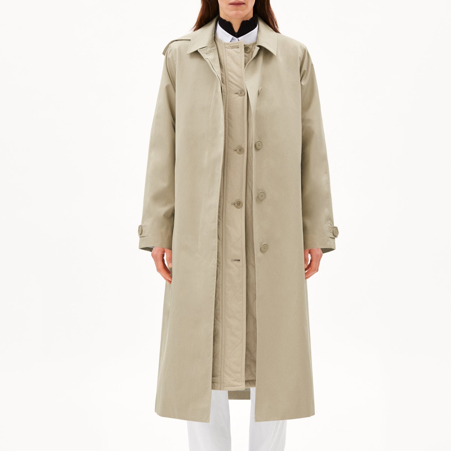 Person in langem, beigem Trenchcoat über weißem Hemd und Hose vor weißem Hintergrund.