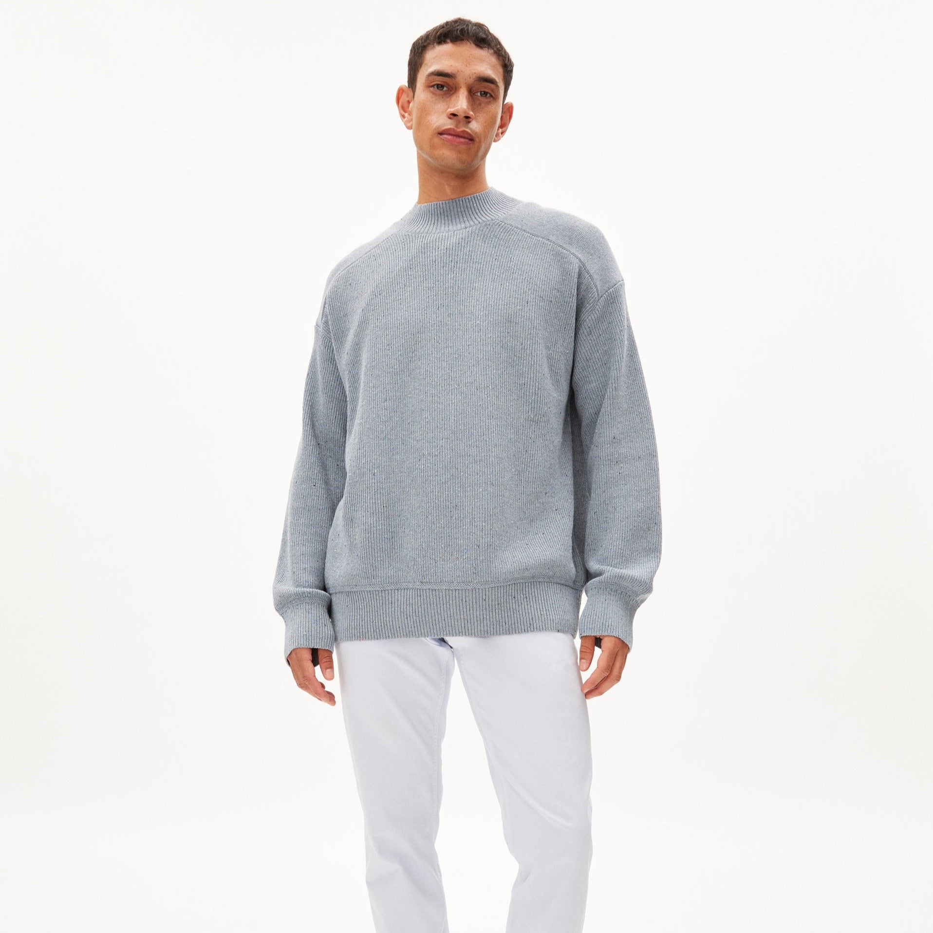 Person in grauem Pullover und weißen Hosen steht vor weißem Hintergrund.