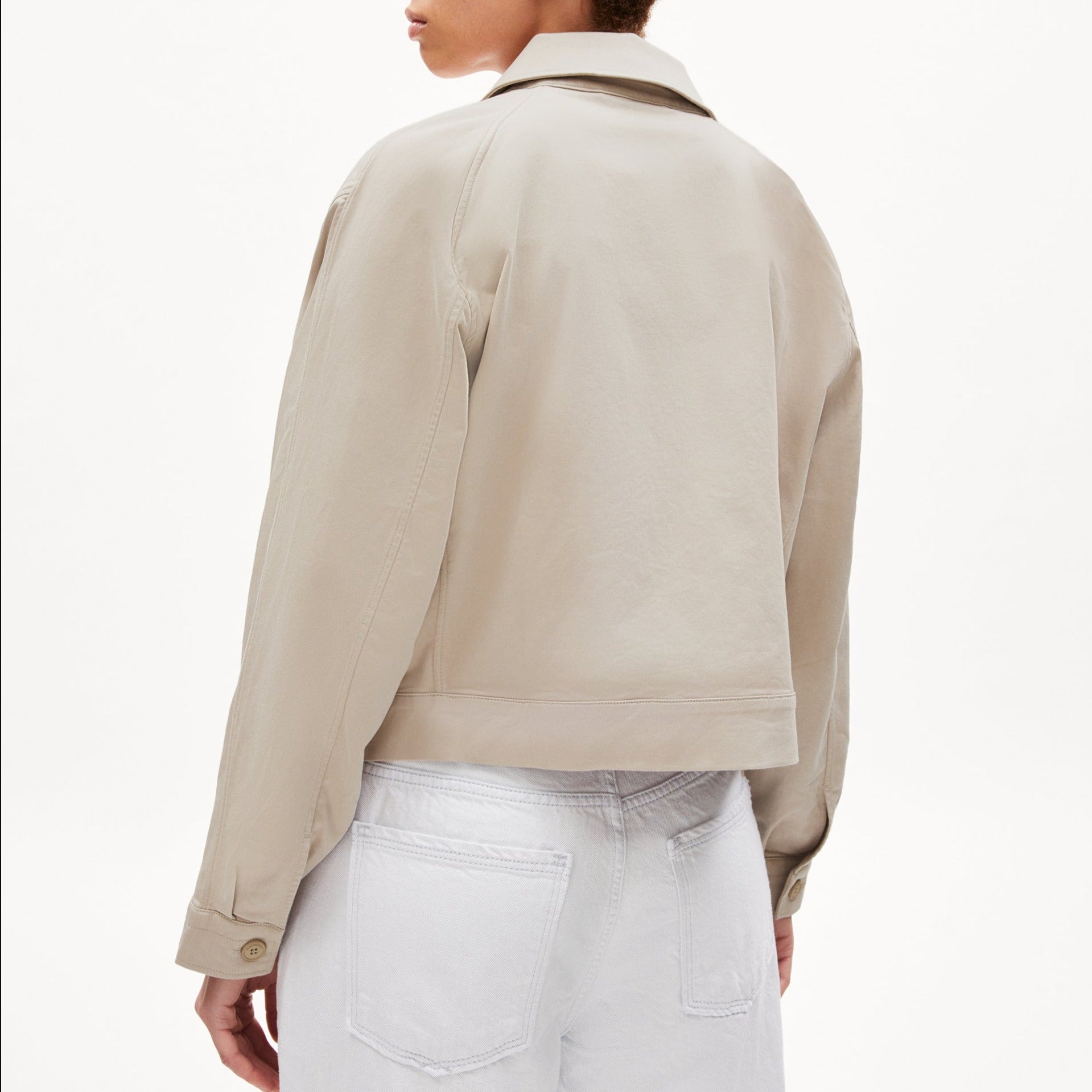 Eine Person trägt eine beige Jacke und weiße Hose, von hinten fotografiert, vor einem weißen Hintergrund.