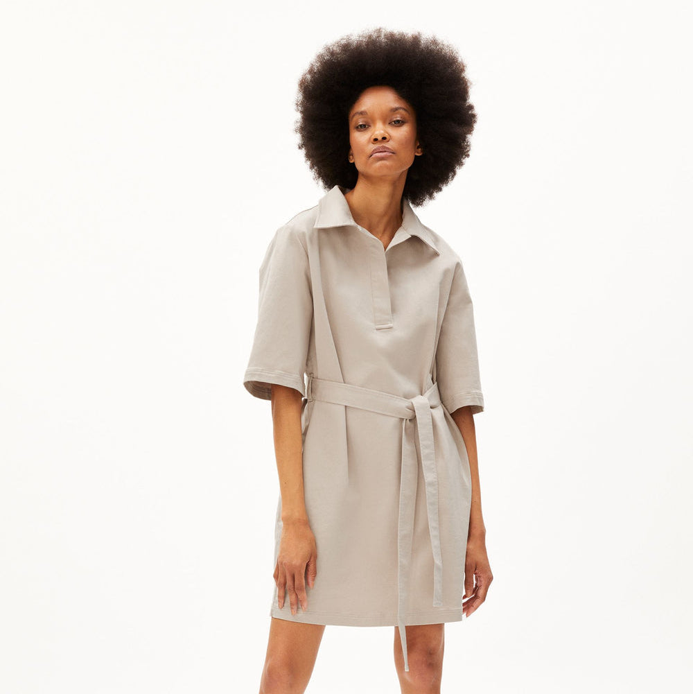 Eine Frau mit Afrofrisur trägt ein beigefarbenes, kurzärmeliges Hemdkleid mit Kragen und einer Krawatte in der Taille, gepaart mit passenden beigen Slippern, vor einem schlichten weißen Hintergrund.