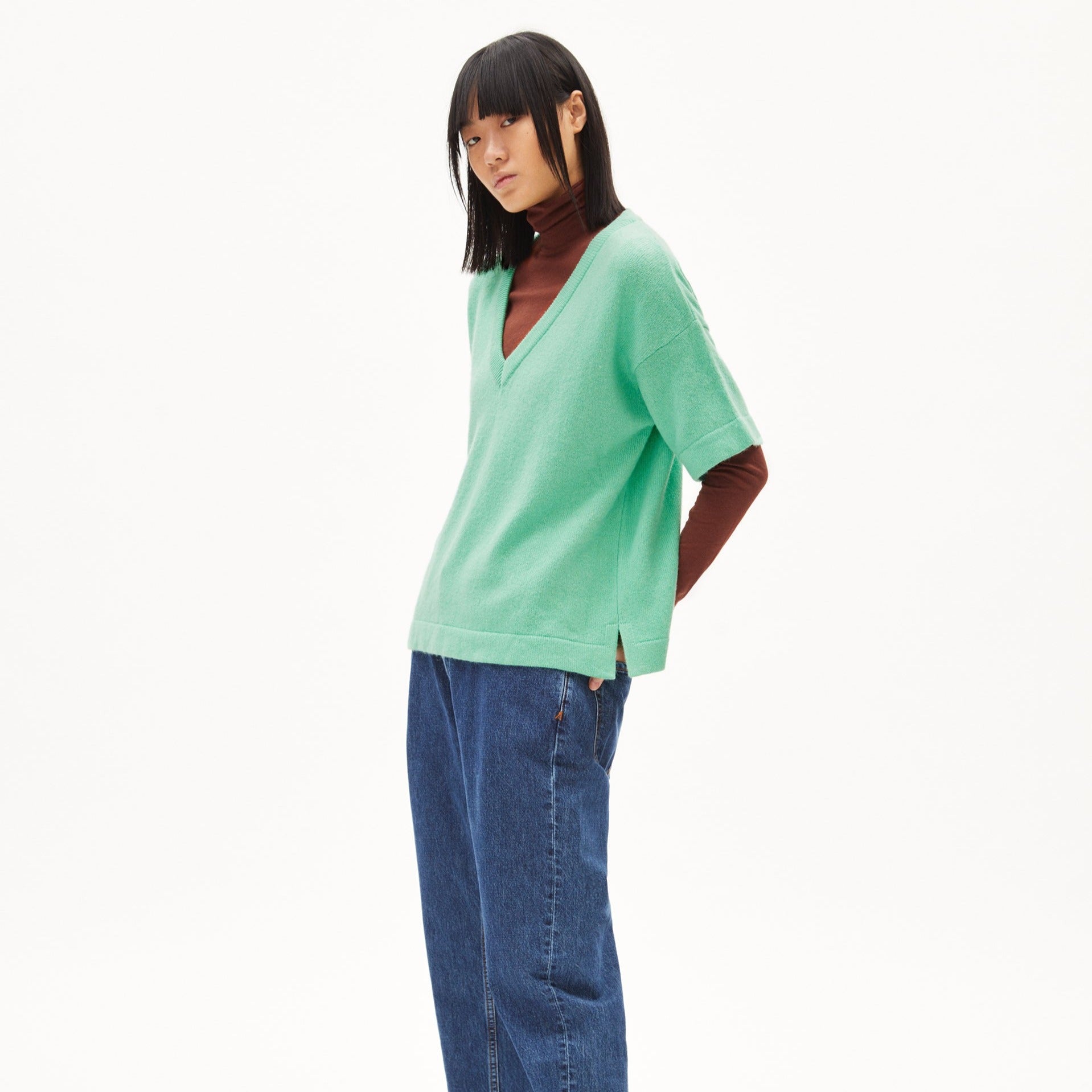 Person in mintgrünem Pullover mit V-Ausschnitt und braunem Rollkragen, trägt blaue Jeans vor weißem Hintergrund.