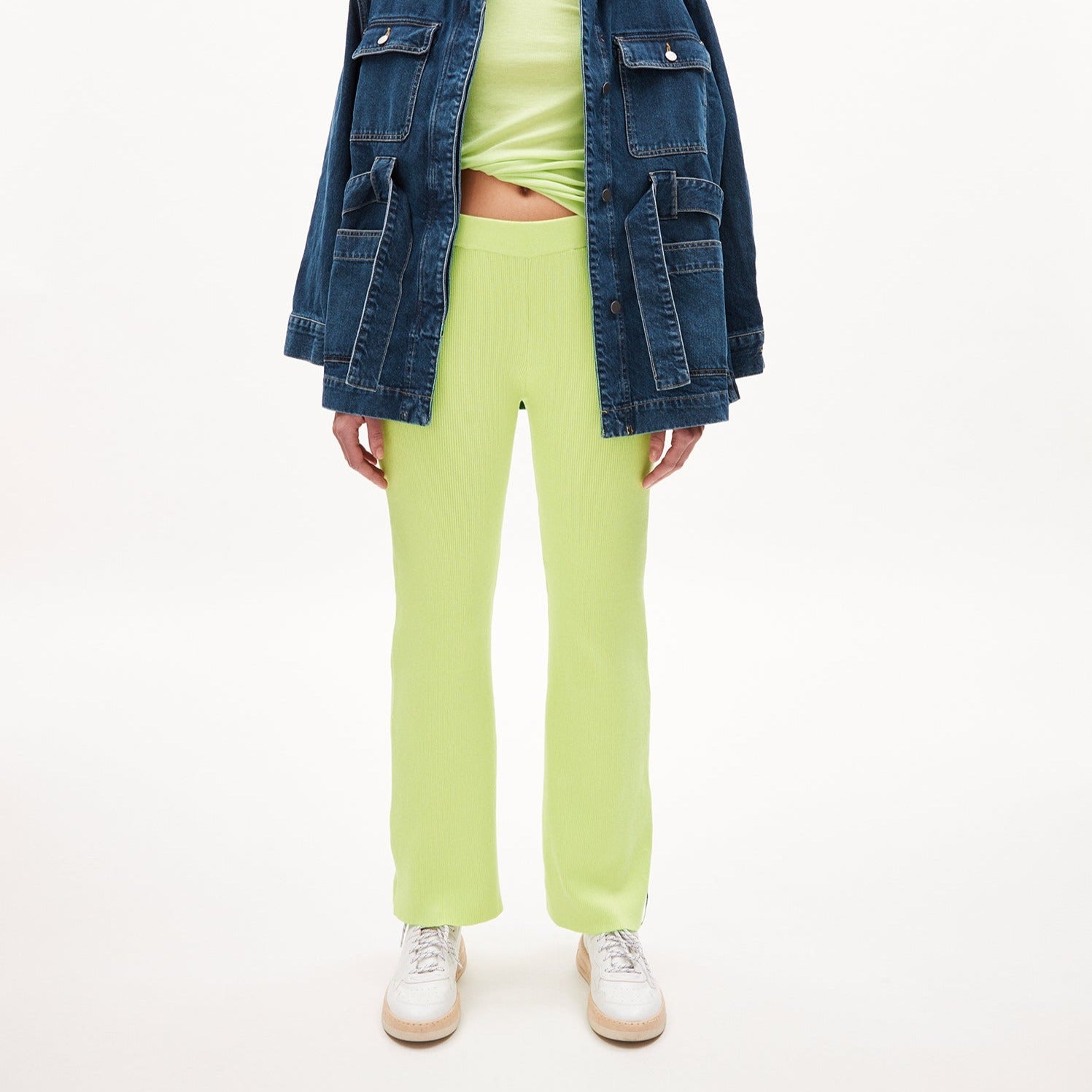 Person in neongrüner Hose, grünem Oberteil und blauer Jeansjacke vor weißem Hintergrund.