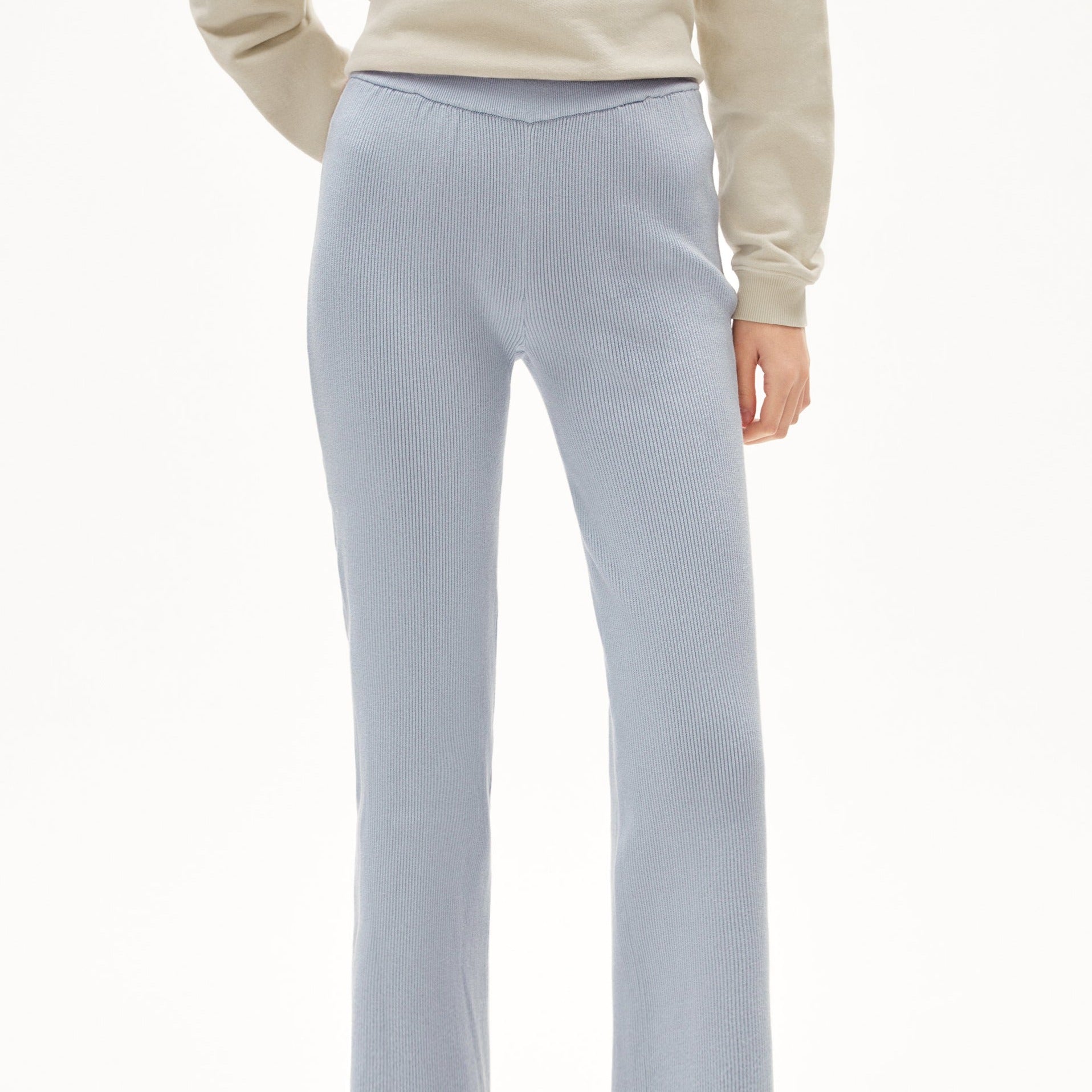 Person in beige Pullover trägt hellblaue, gerippte Hose. Nur Unterkörper und Hände sichtbar. Hintergrund ist weiß.