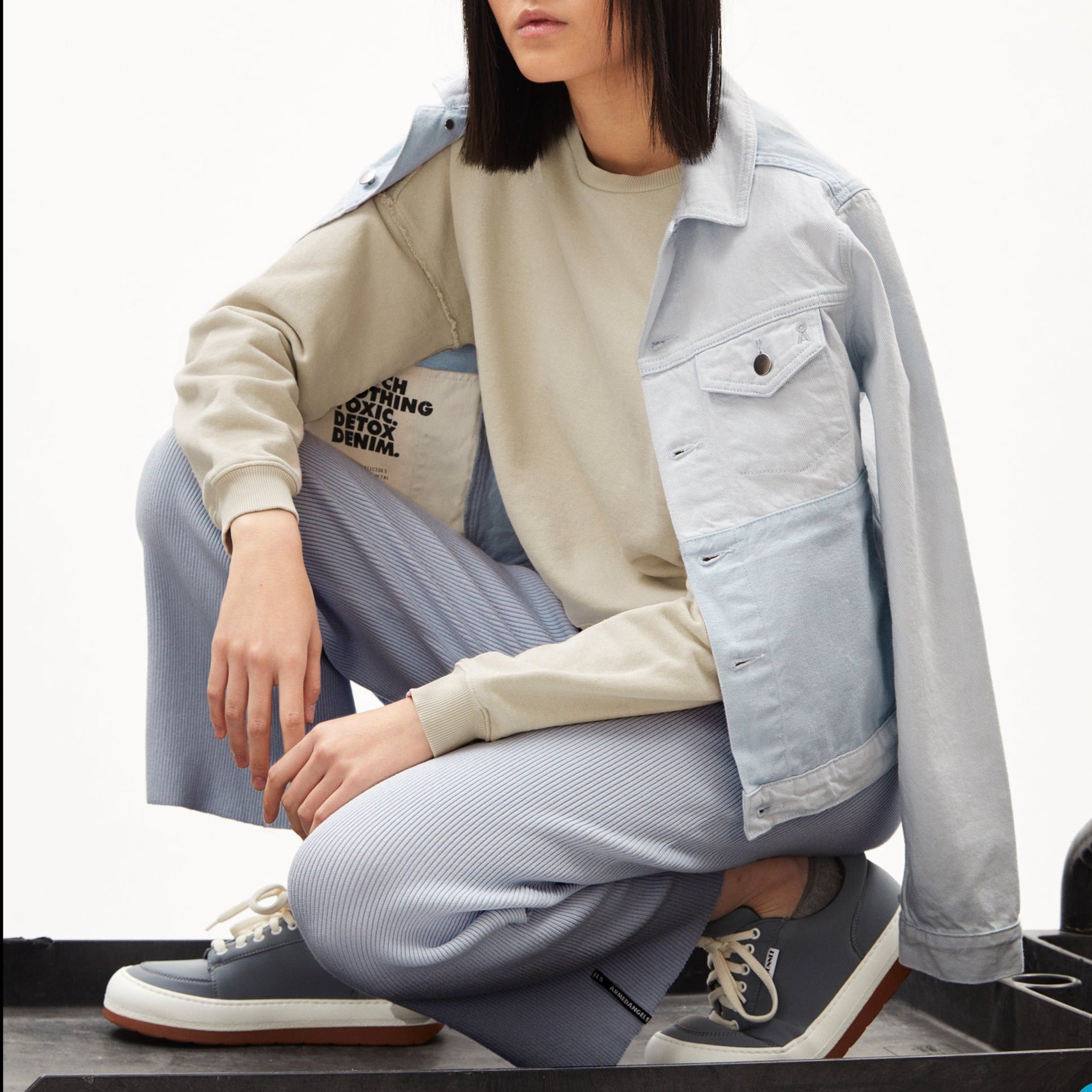 Person in hellem Sweatshirt und Jeansjacke kniet auf einem Bein, trägt blaue Hosen und graue Turnschuhe.
