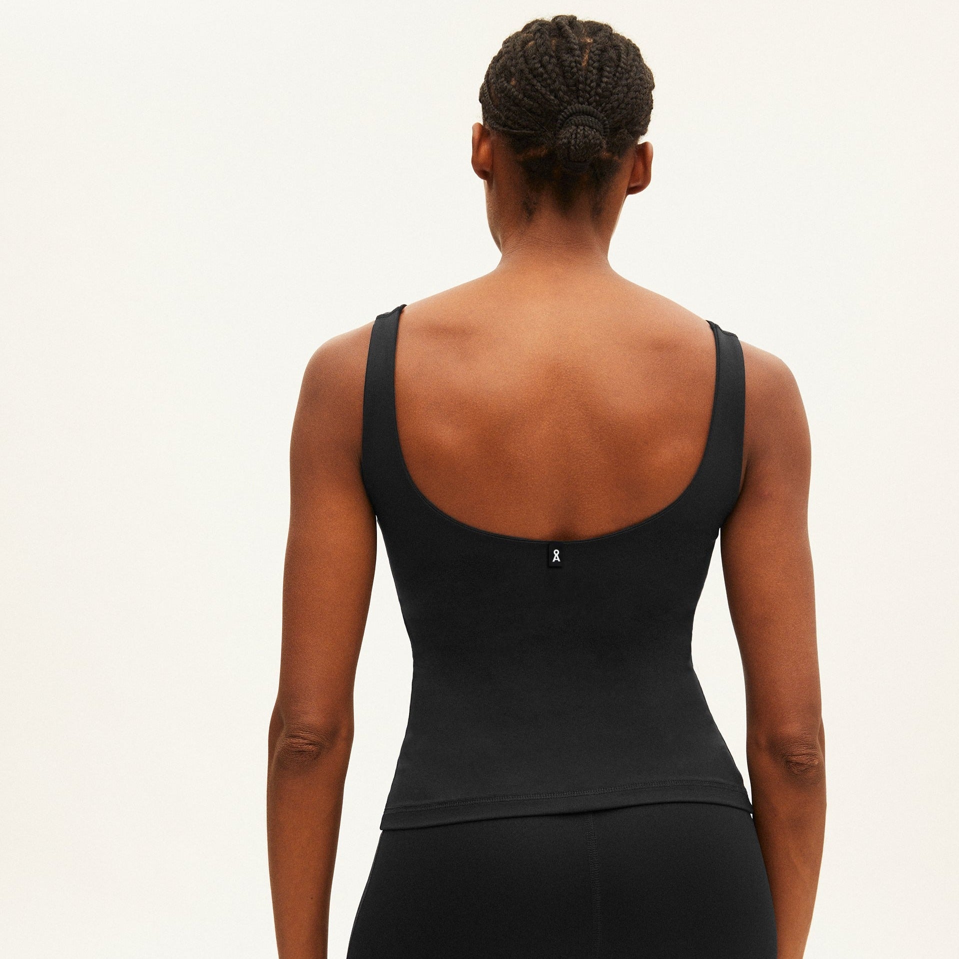 Person in schwarzem Top von hinten, Haare in geflochtenen Zöpfen, vor einem hellen Hintergrund.