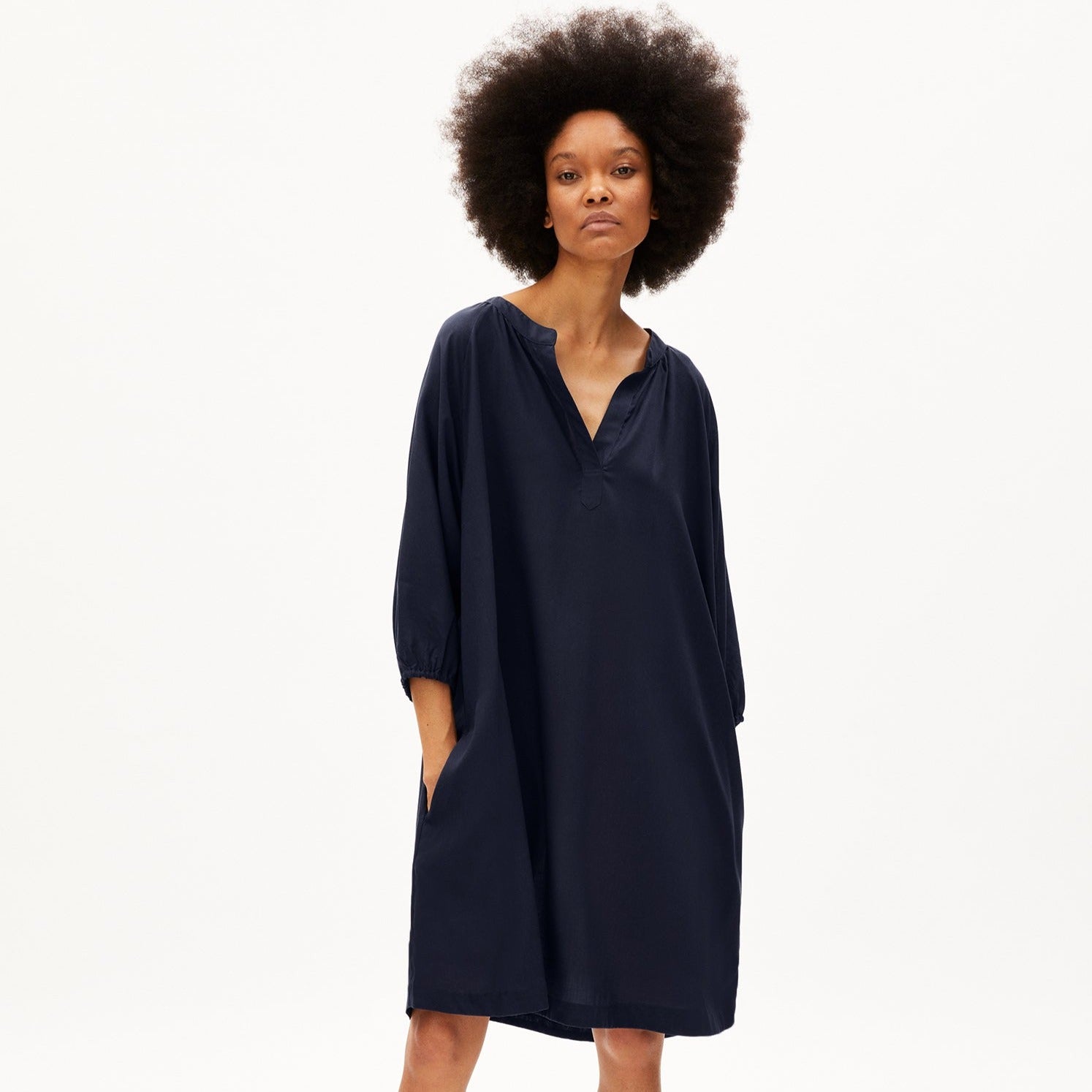 Person in lockerem, dunkelblauem Kleid mit dreiviertel Ärmeln und lockigem Haar vor weißem Hintergrund.