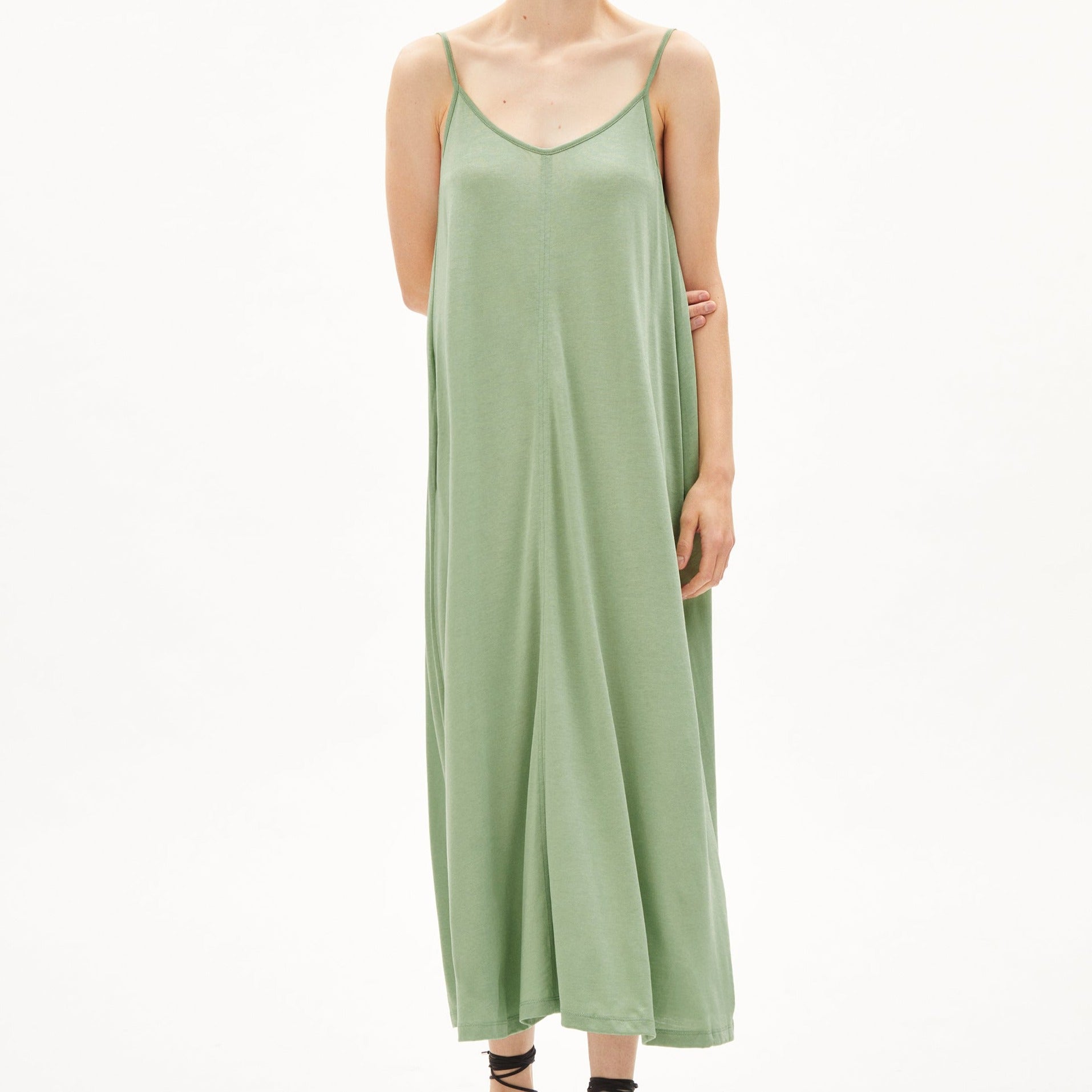 Person in langem, grünem Maxikleid mit Spaghettiträgern steht vor einem weißen Hintergrund.