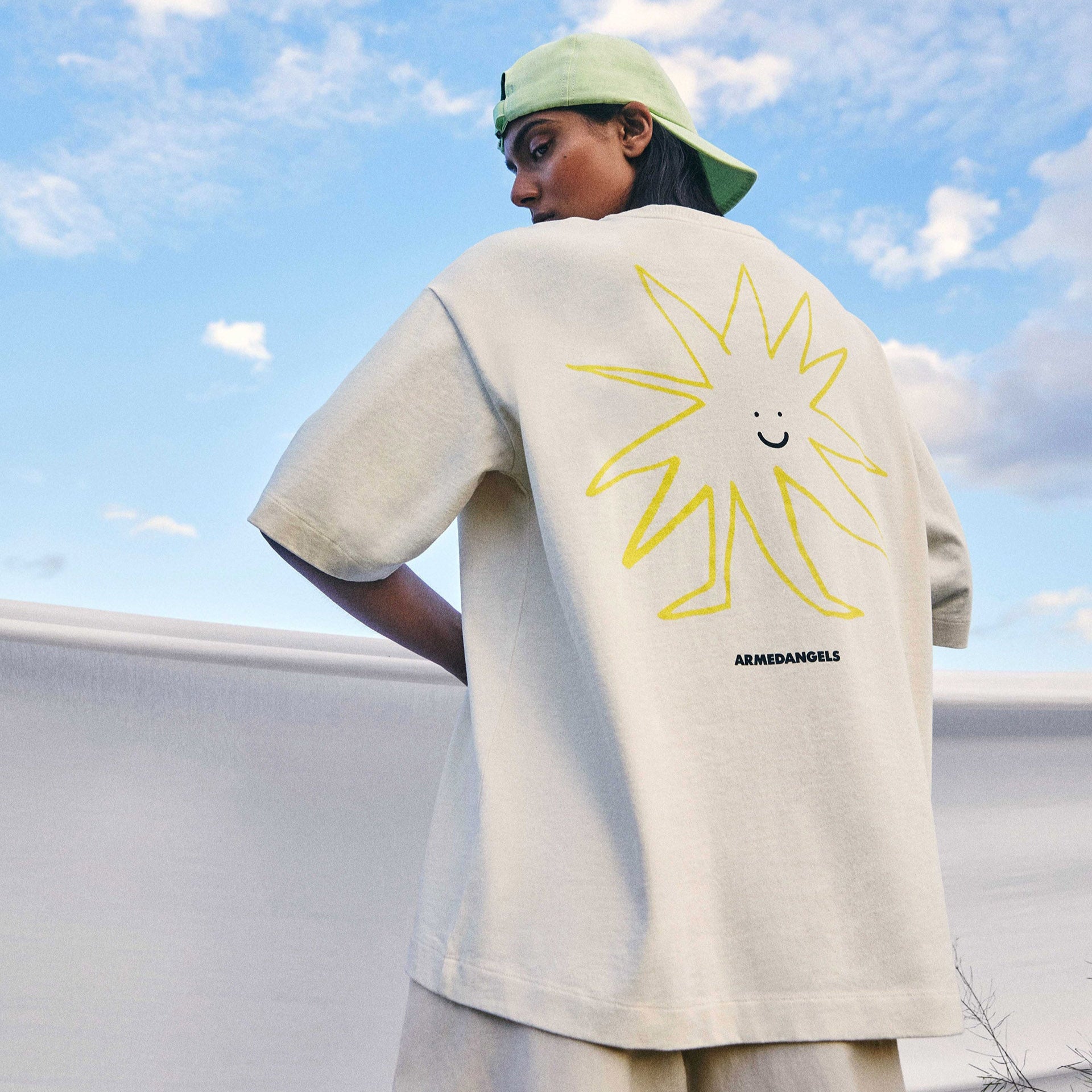 Person trägt ein cremefarbenes Shirt mit gelbem Sonne-Design, steht vor einer blauen Himmelkulisse.